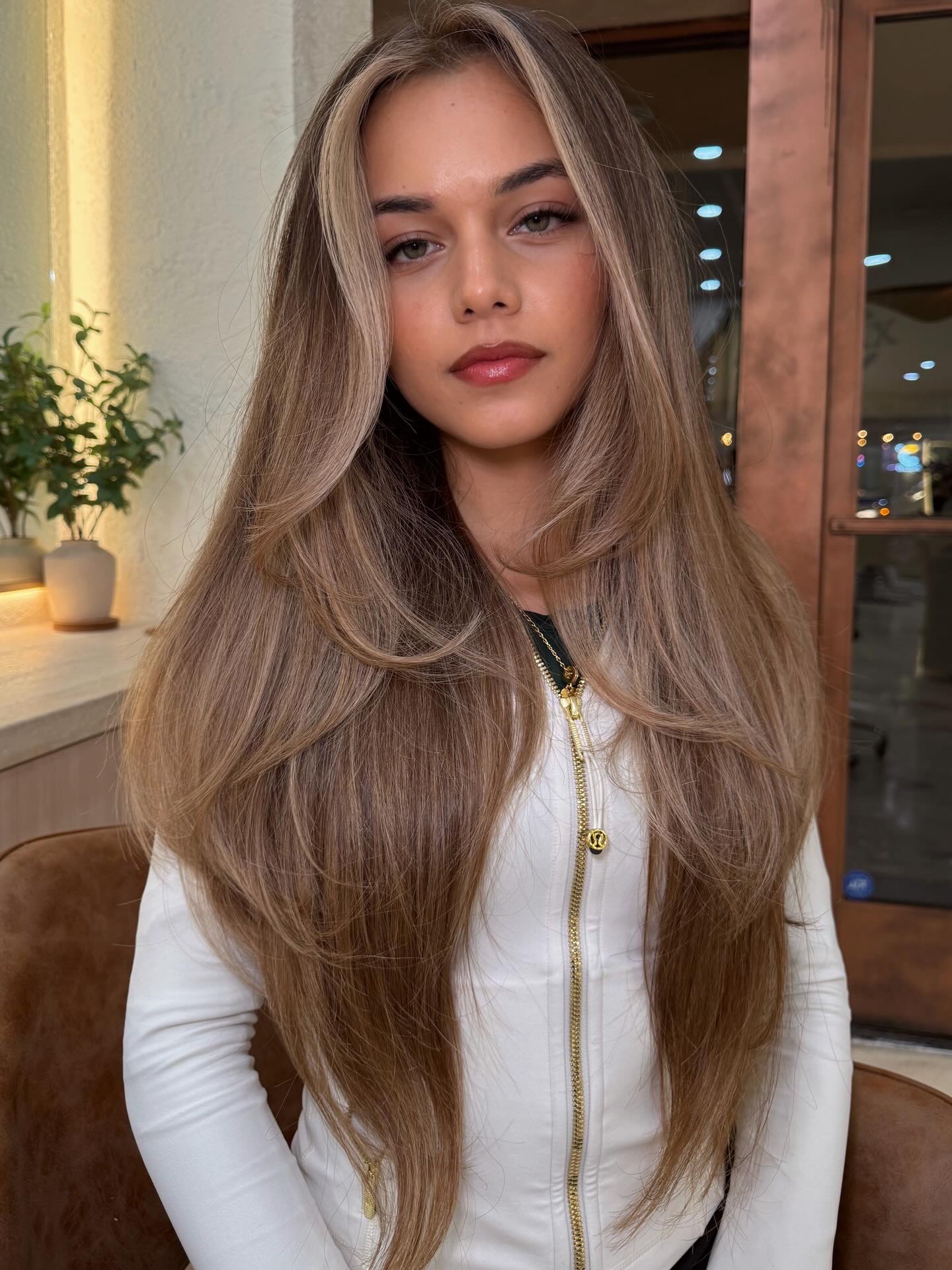 Young woman with long, wavy hair and neutral expression, wearing a white zip-up jacket, indoors with warm lighting.