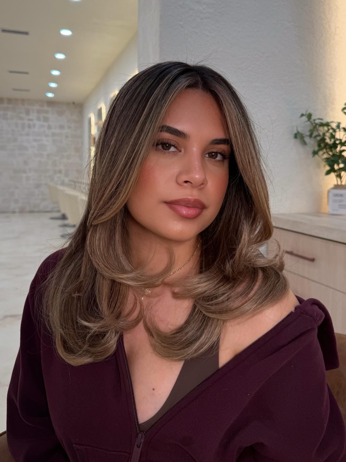 Young woman with shoulder-length hair and light makeup, wearing a dark top, in an indoor setting with plants and lighting.