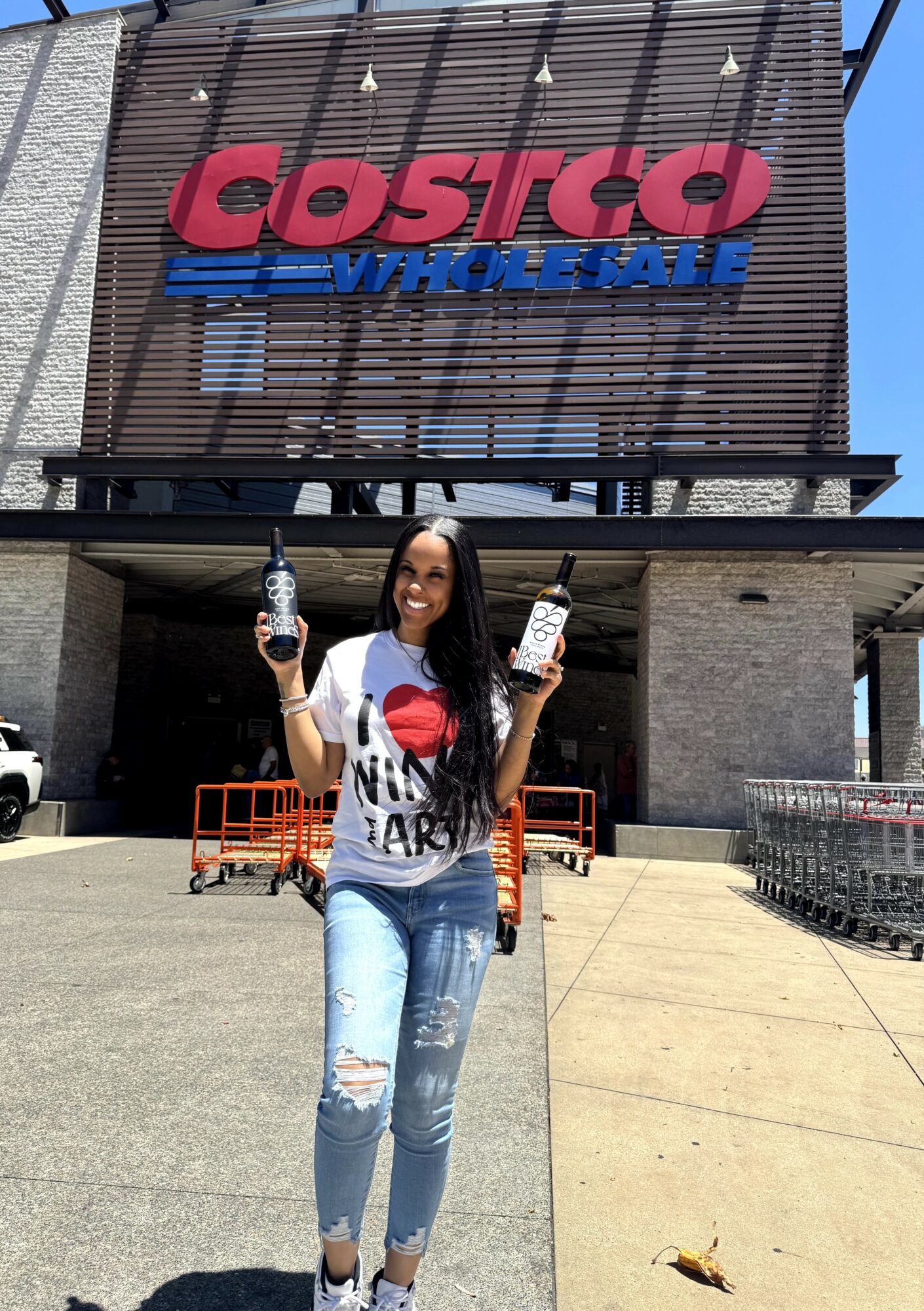 Woman holding two bottles in front of Costco Wholesale store entrance, smiling, wearing a white T-shirt and ripped jeans.