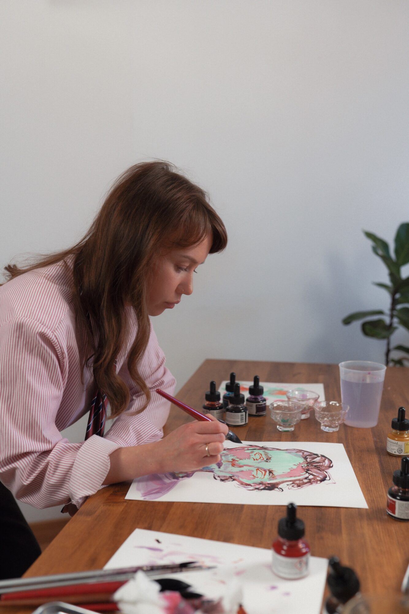 Woman with long brown hair painting at a table with art supplies, a plant, and a glass of water nearby.