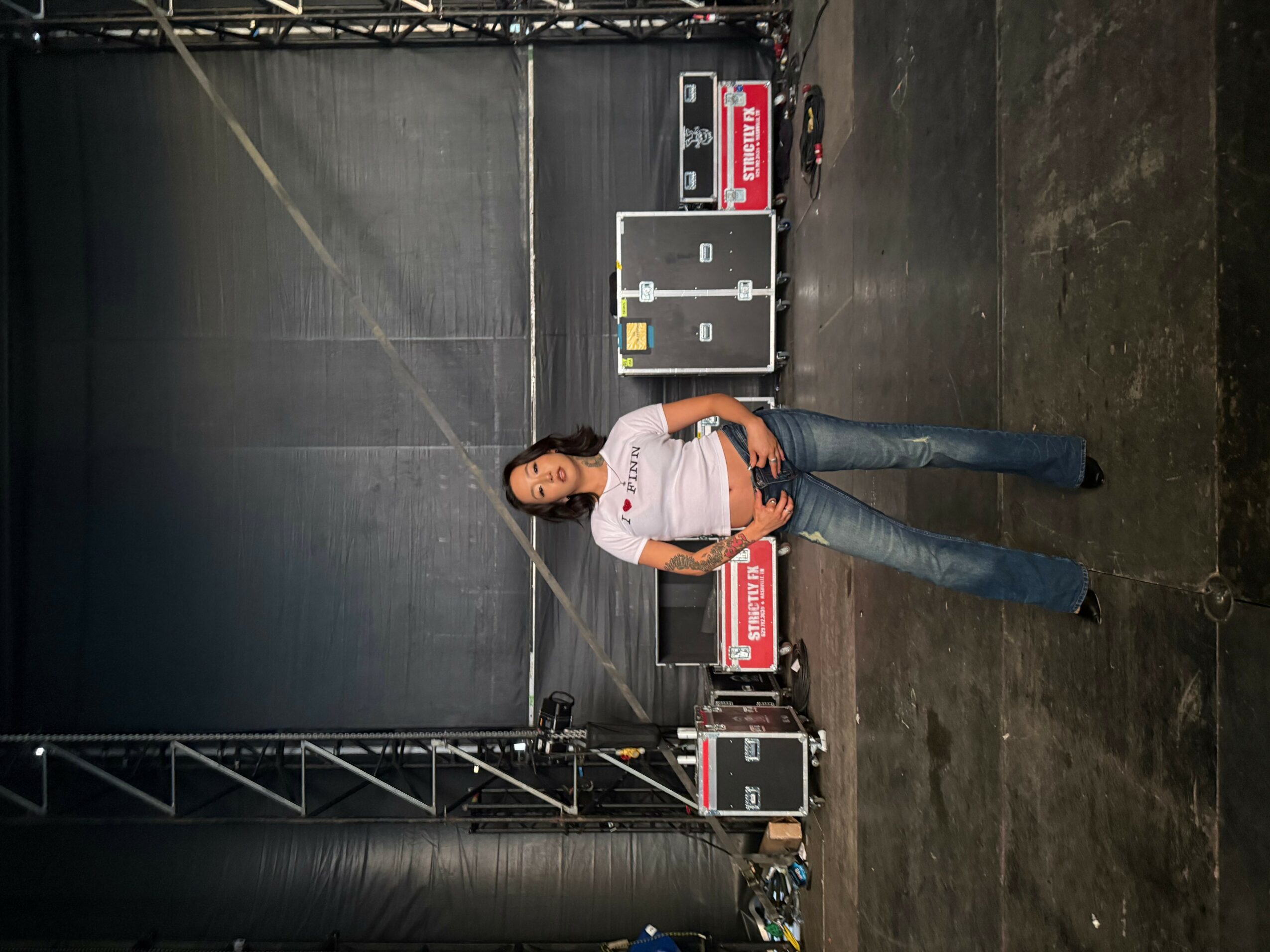 Woman standing in front of a black industrial door, wearing a white shirt and jeans, with equipment and signs nearby.