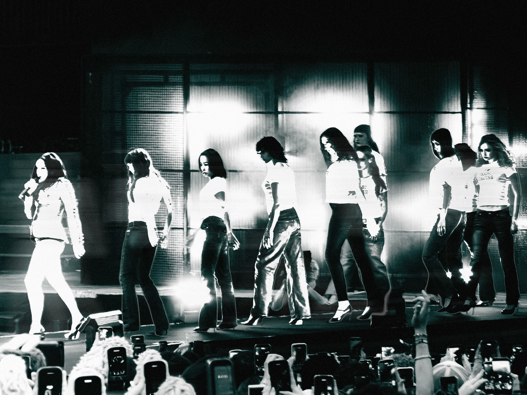 Group of people walking on stage with bright backlighting, audience taking photos, in black and white.