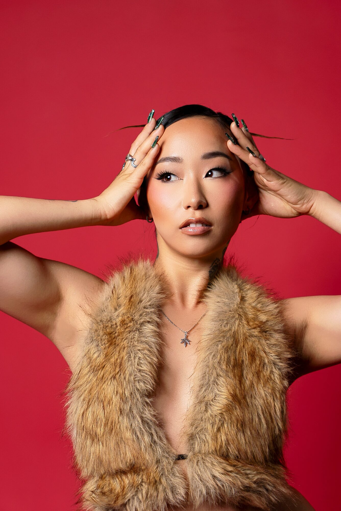 Woman with hands on head, wearing fur vest, against red background, looking to the side.