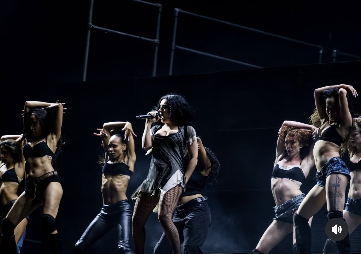 Group of women performing on stage, some with hands on their heads, dark background, stage lighting, energetic scene.