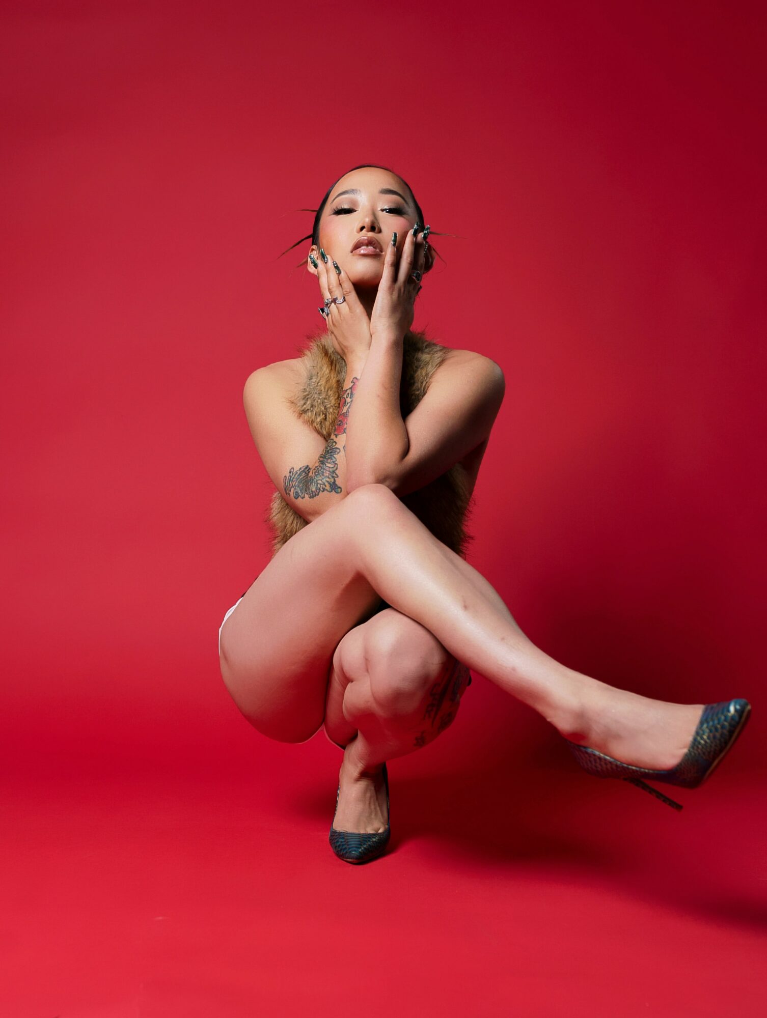 Woman with tattoos poses with one leg crossed over the other against a red background, touching her face with one hand.