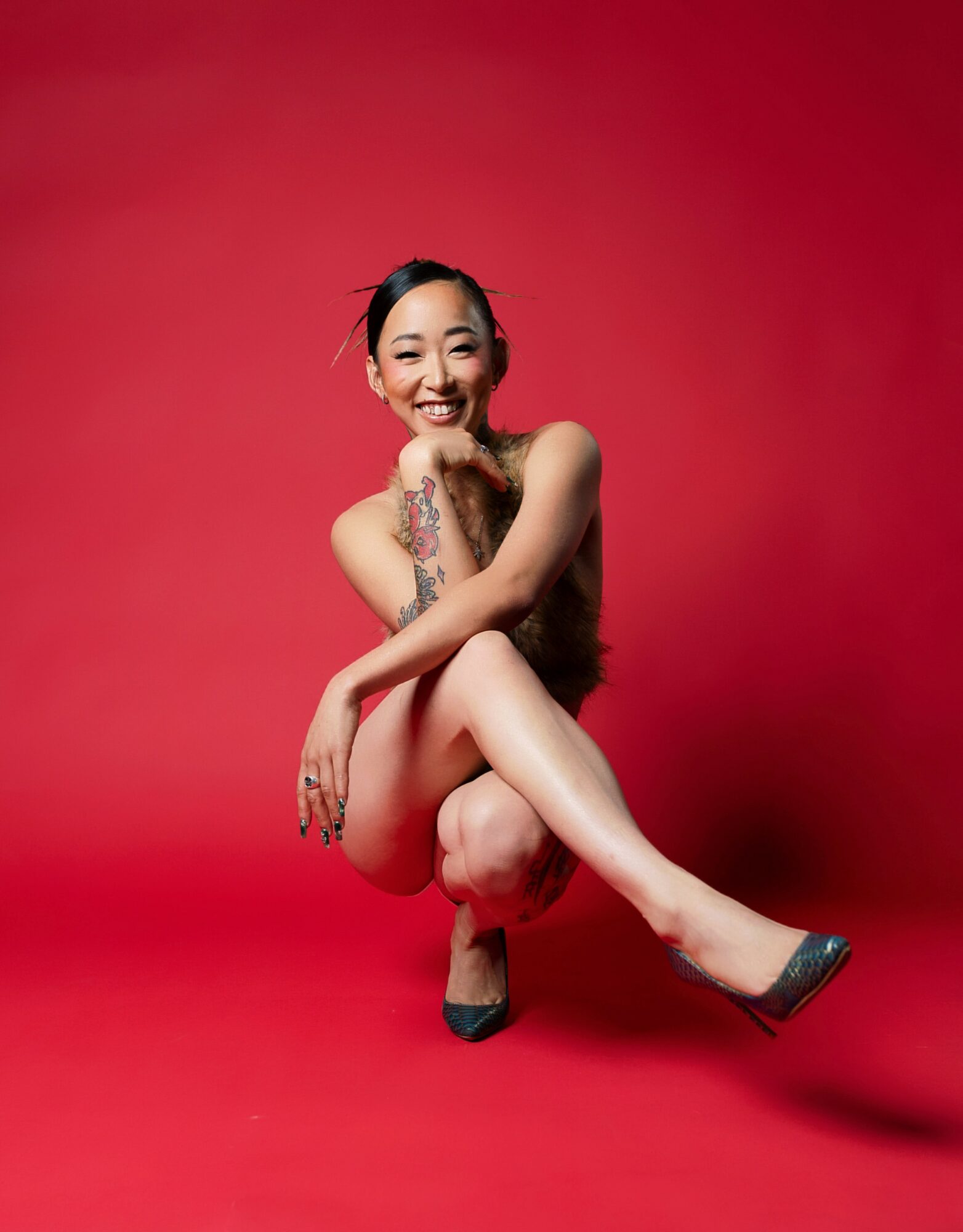 Smiling woman with tattoos, sitting with one leg crossed over the other, against a red background.