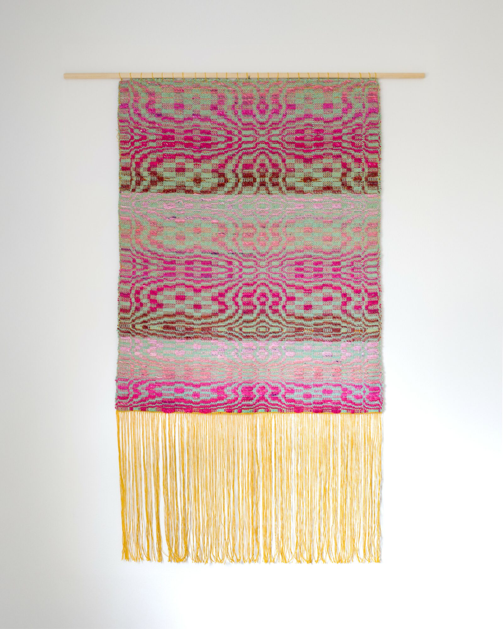 Colorful woven wall hanging with pink, beige, and brown patterns, and long yellow fringe at the bottom.
