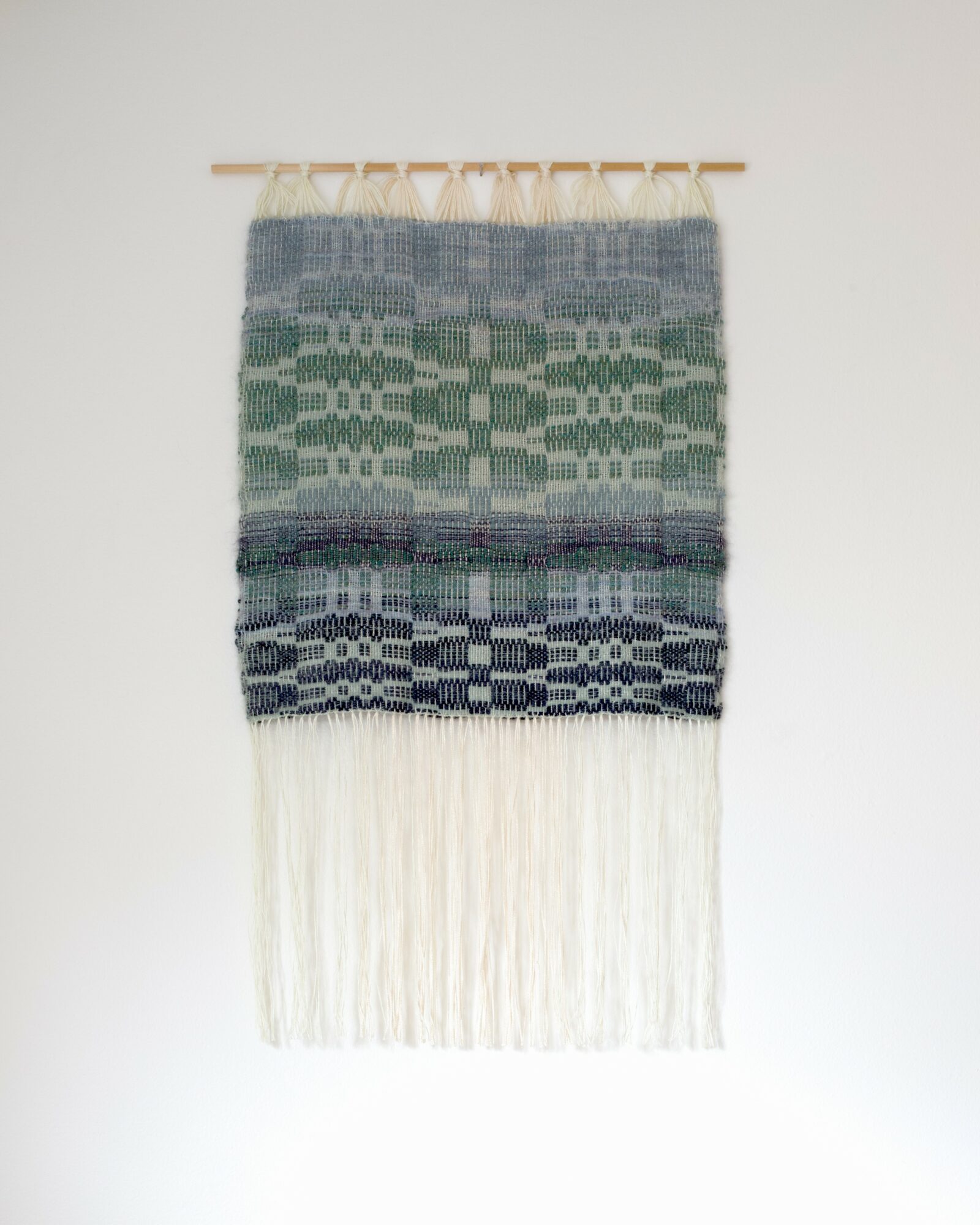Textured woven wall hanging with horizontal bands of green, blue, and white, and long white fringe at the bottom.