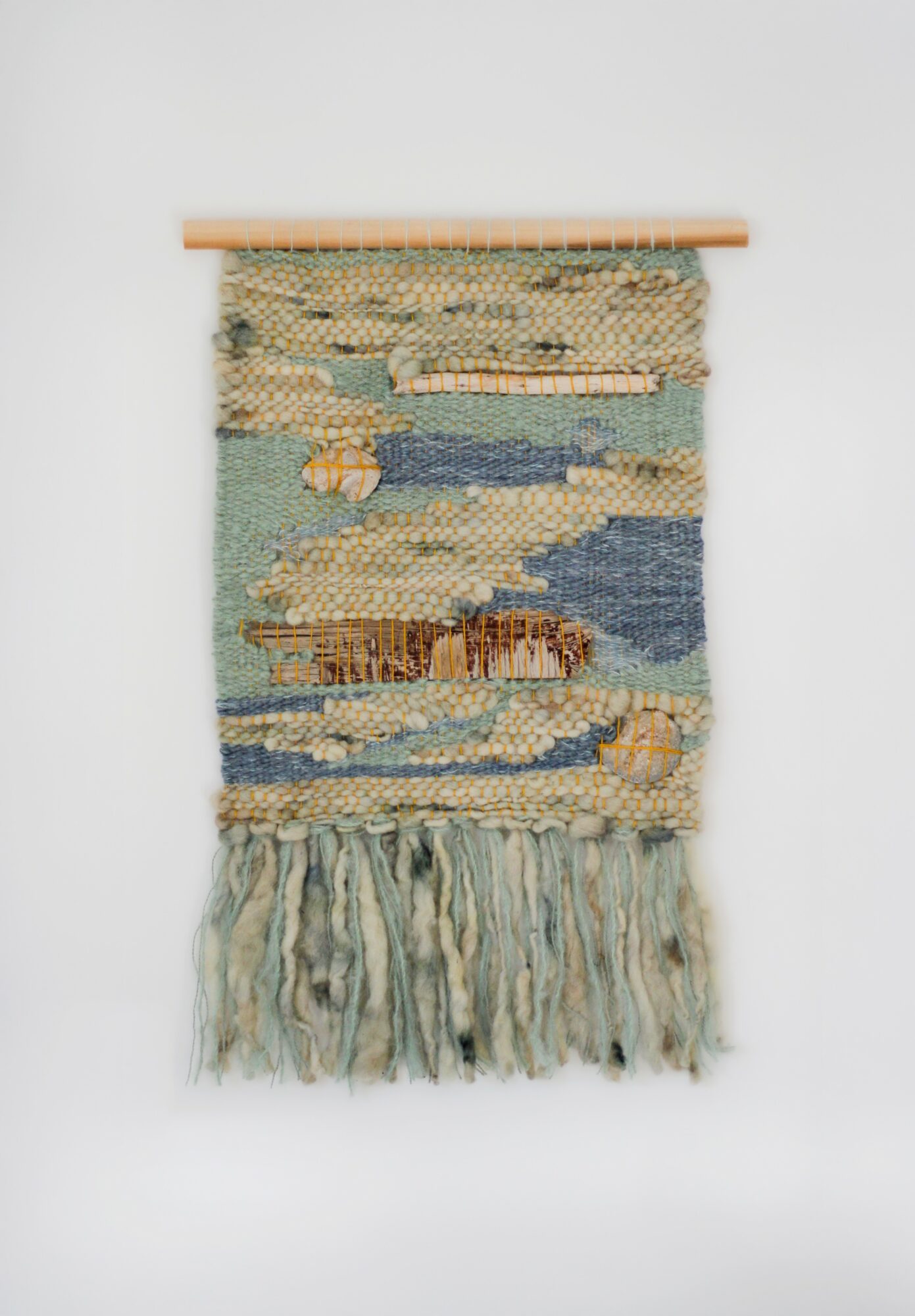 Textured woven wall hanging with landscape scene, hanging from a wooden rod, with fringes at the bottom.