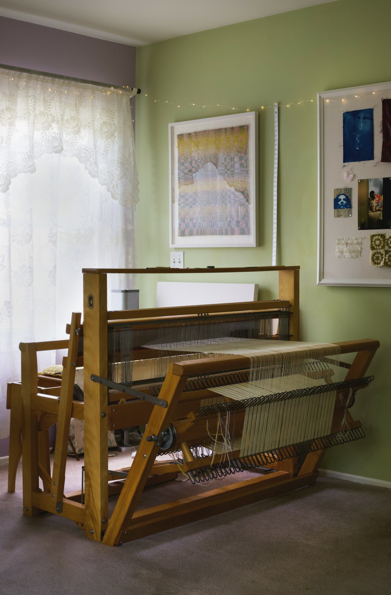 Wooden drying rack with woven surface in a room with green walls and framed artwork, near a window with lace curtains.