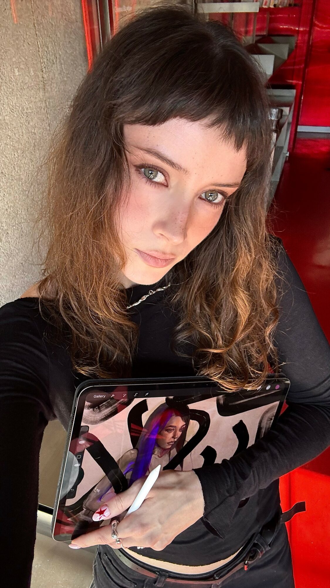 Young woman with wavy brown hair and bangs, holding a tablet, looking at the camera, in a red and black environment.
