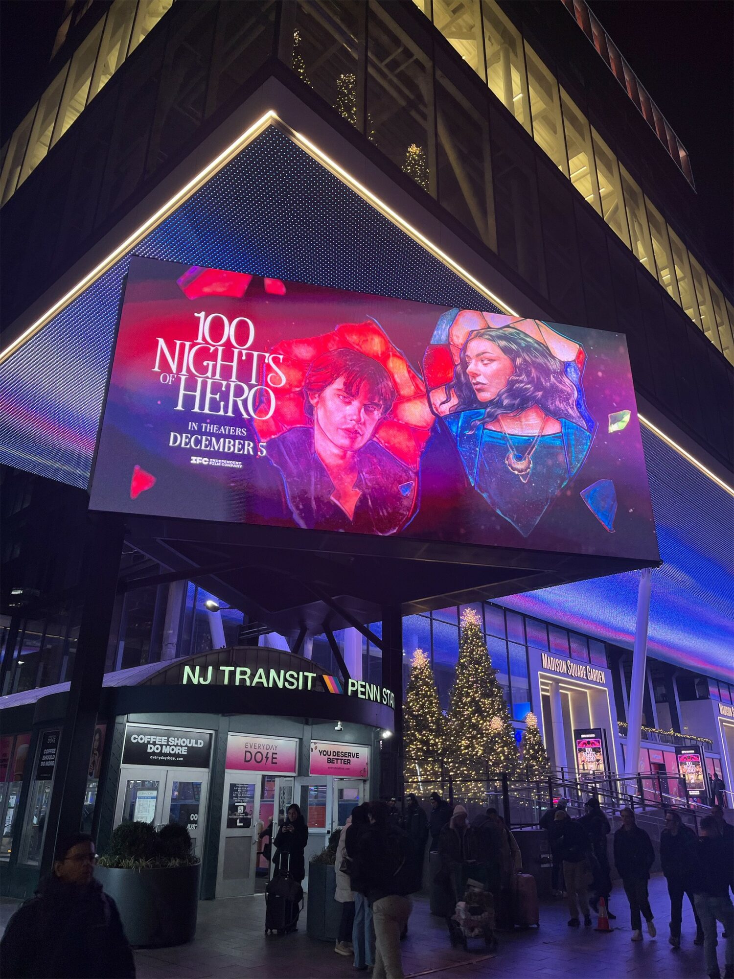 Large digital billboard displays an advertisement for a music event with two women, city street scene below, people walking, nighttime setting.