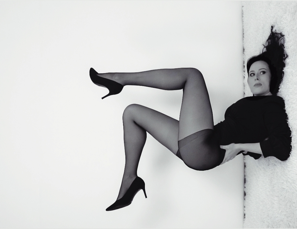 Woman lying on her side with legs raised, wearing black stockings and high heels, against a plain background.