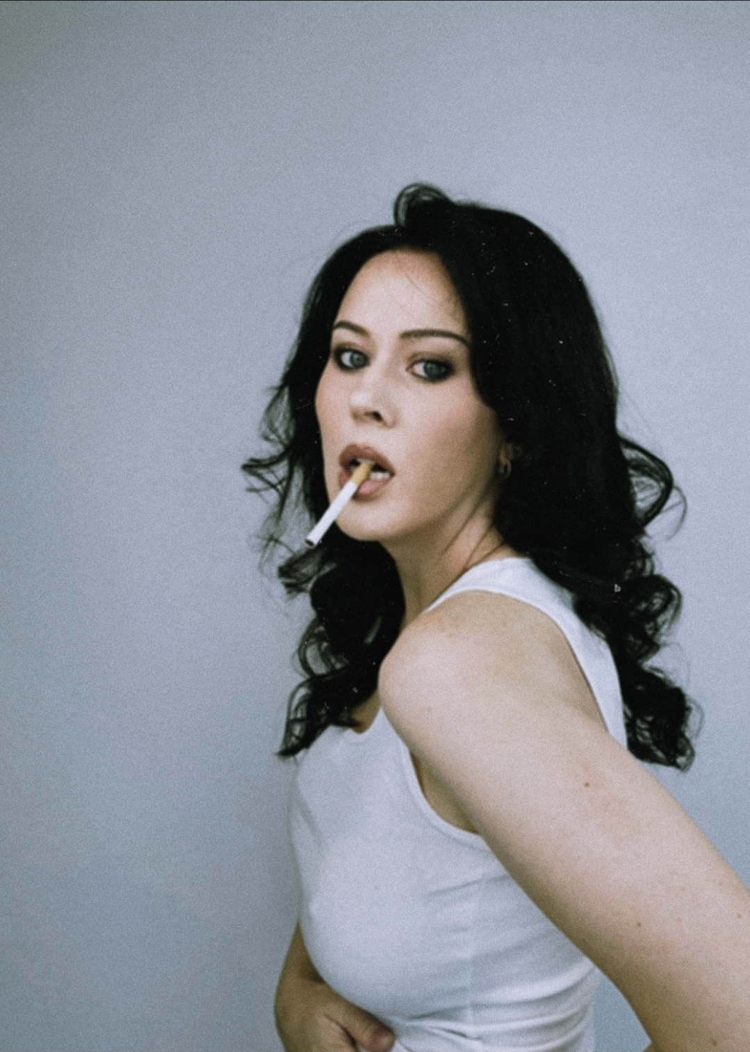 Woman with dark hair and makeup, wearing a white sleeveless top, holding a cigarette in her mouth, looking over shoulder.