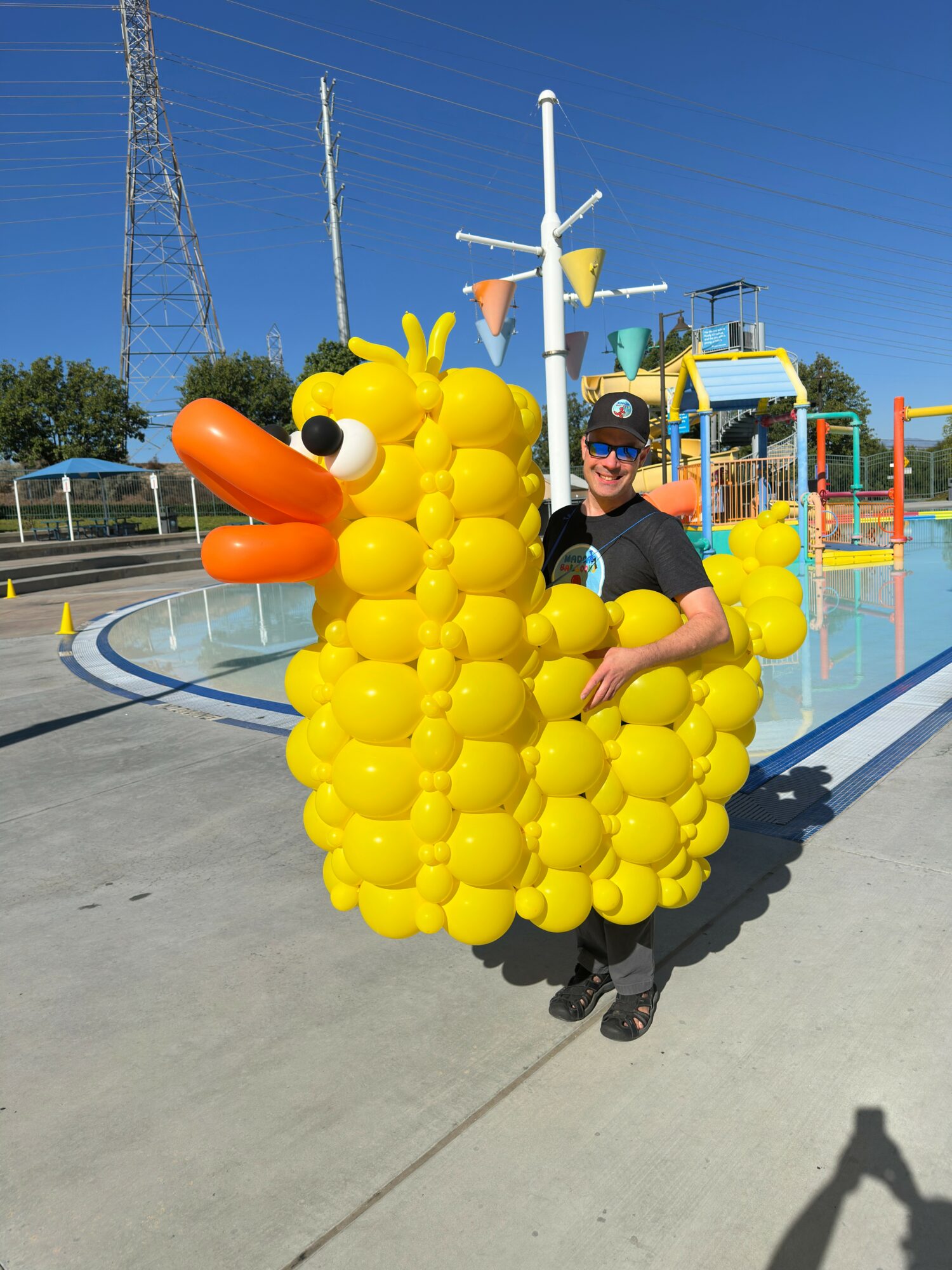 Person standing next to a large yellow duck-shaped balloon sculpture at a water park, with water slides in the background.