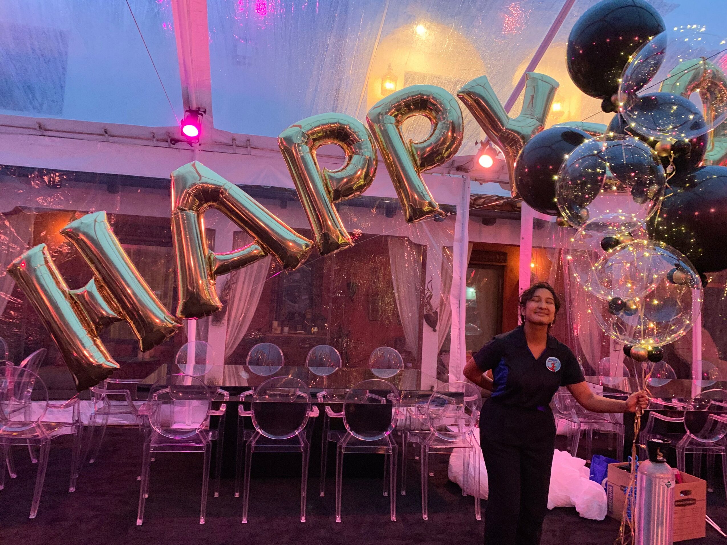 Person standing in decorated party tent with balloons and 'HAPPY' balloons, pink and purple lighting, and chairs in background.