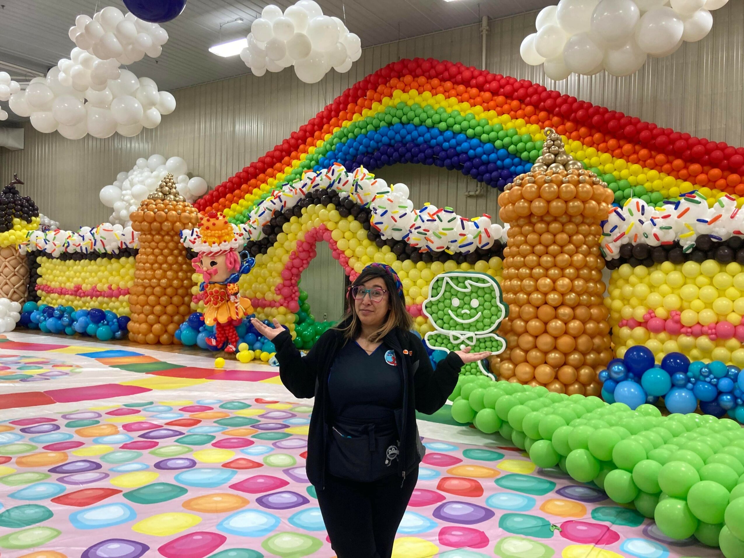 Woman standing in front of colorful balloon rainbow and cloud decorations at an indoor event.
