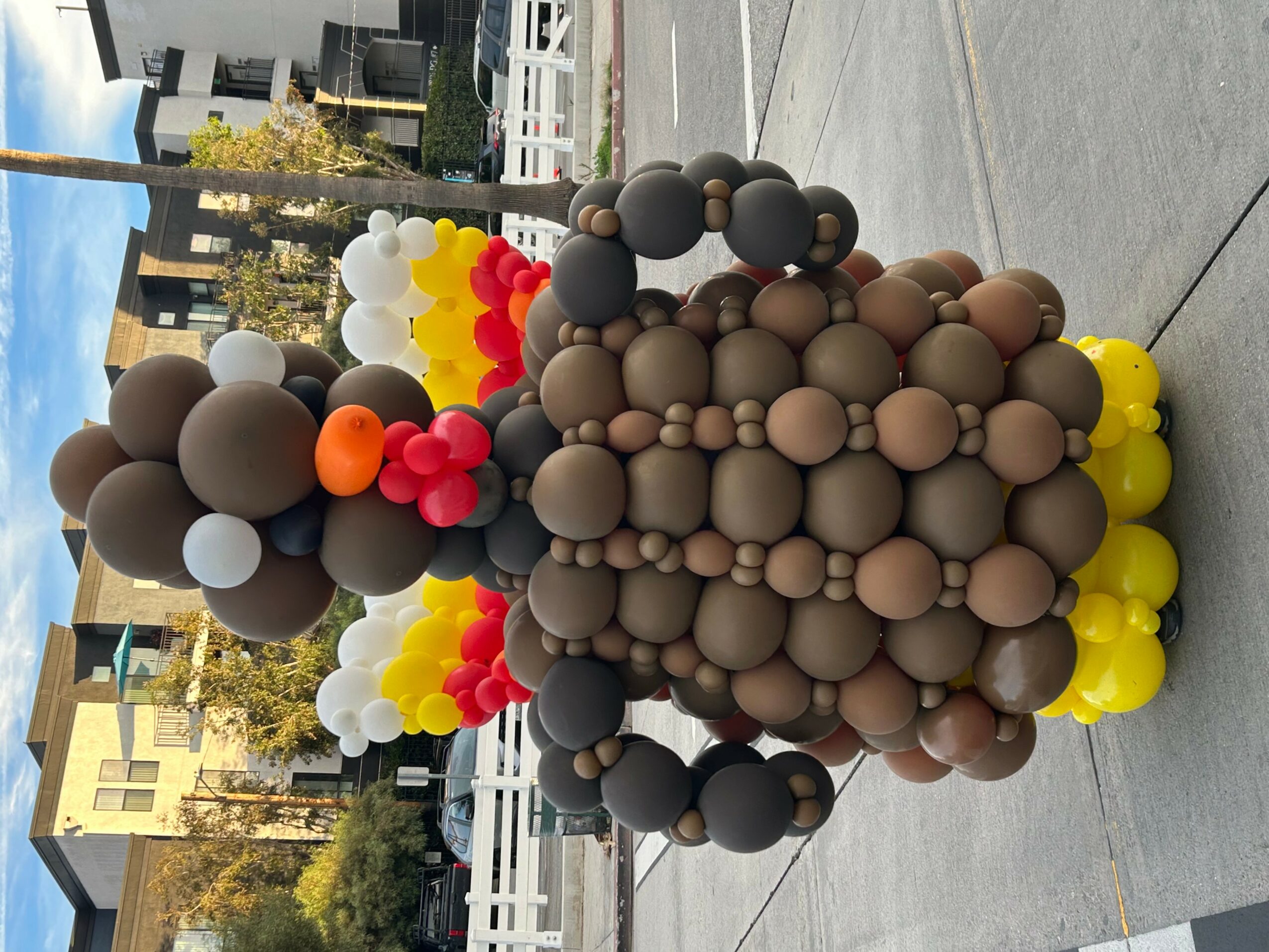 Colorful balloon sculpture resembling a bear with yellow, red, black, and brown balloons, outdoors on a sidewalk.