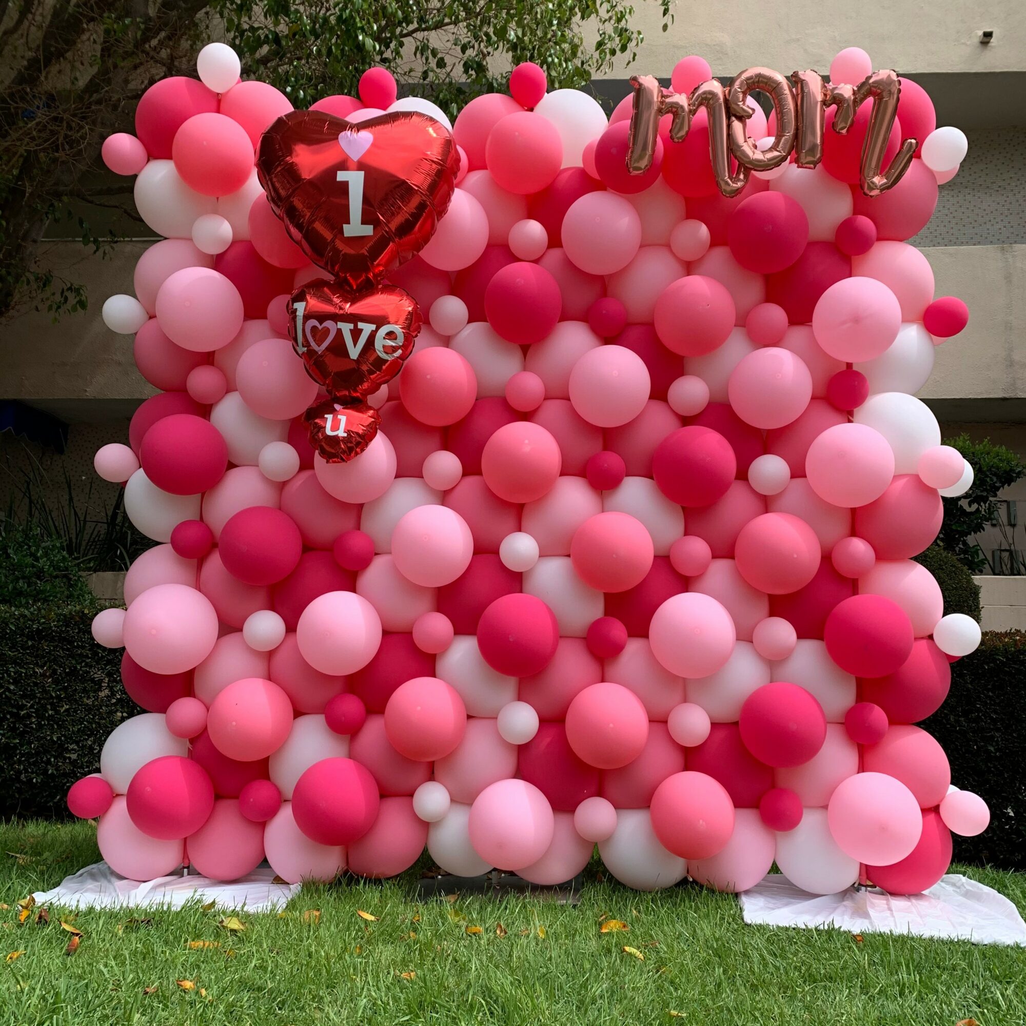 Balloon backdrop with pink, red, and white balloons, heart-shaped balloons, and a pink cloth at the bottom, outdoors.