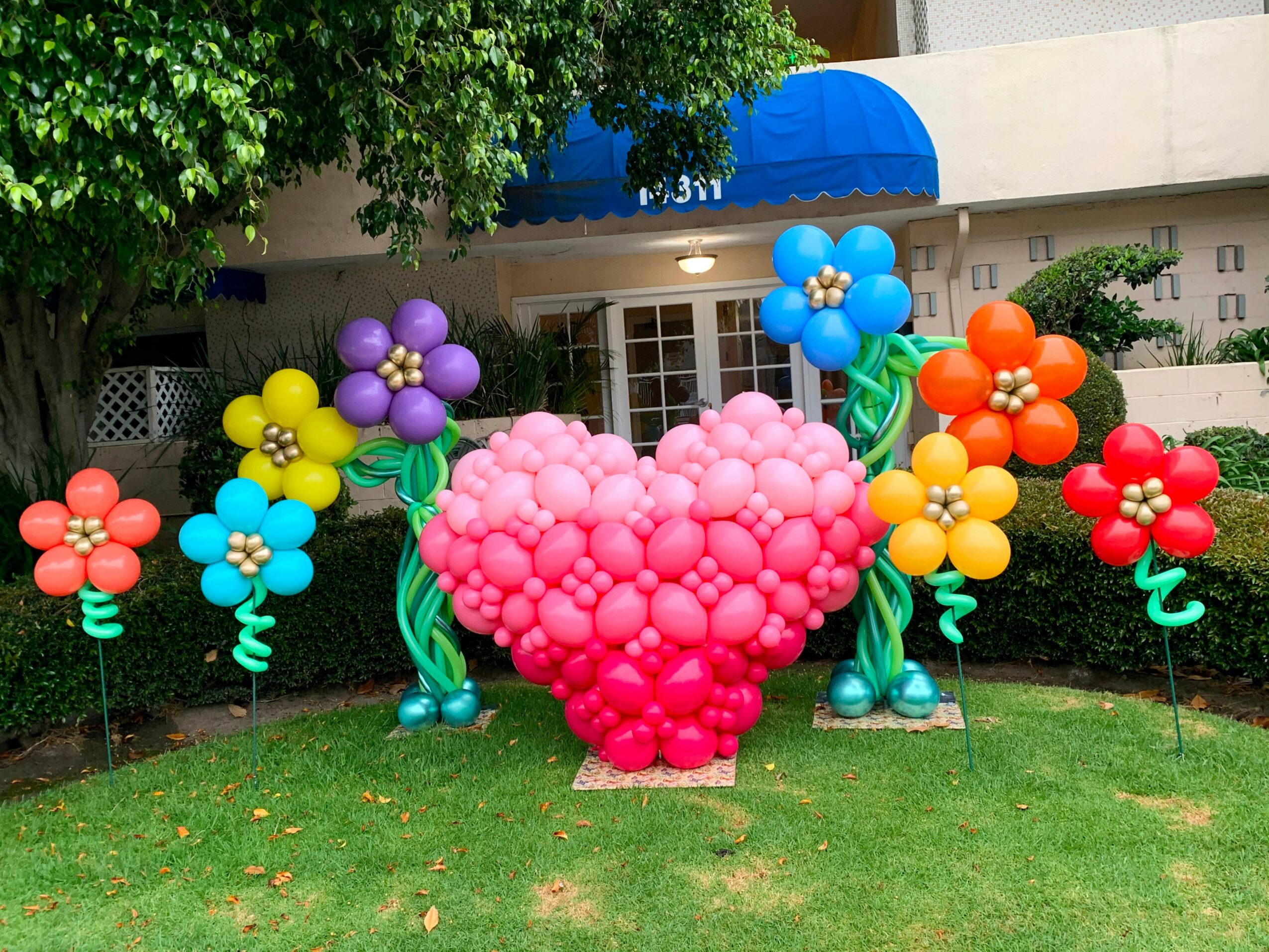 Colorful balloon display with flowers and a heart shape in front of a building, under a tree, on a lawn.