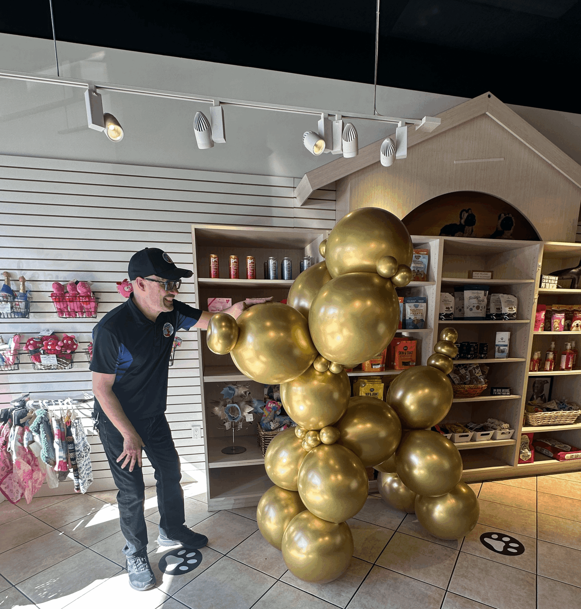 Person wearing black cap and glasses looking at a cluster of large gold balloons inside a store, shelves with various items in background.