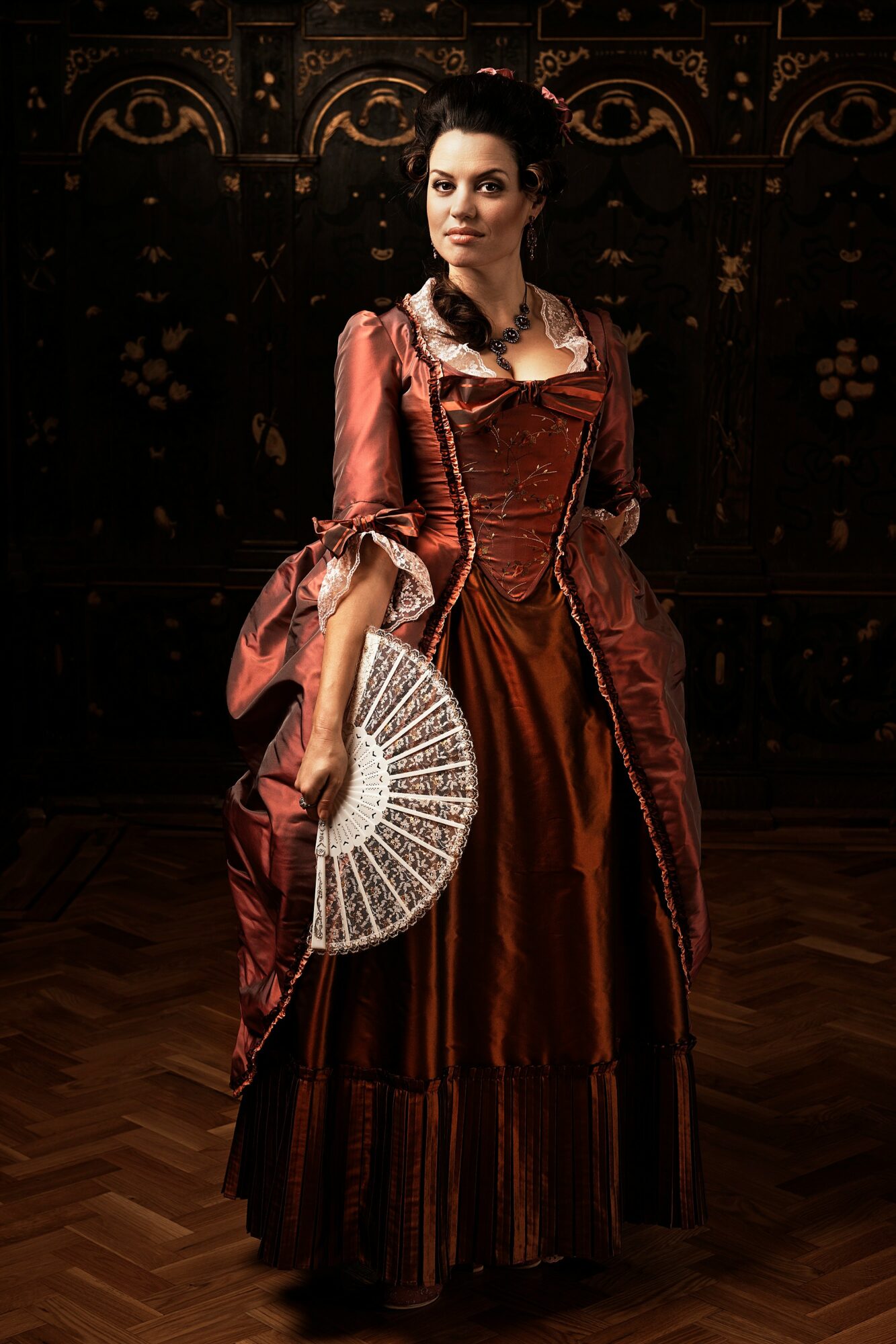 Woman in historical dress holding a fan, standing in a dimly lit room with ornate decor.