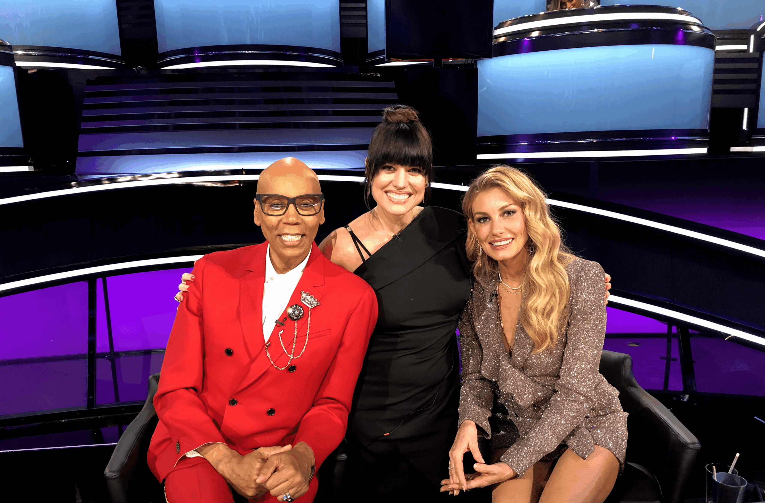 Three women sitting together on a stage with colorful lighting, smiling at the camera.