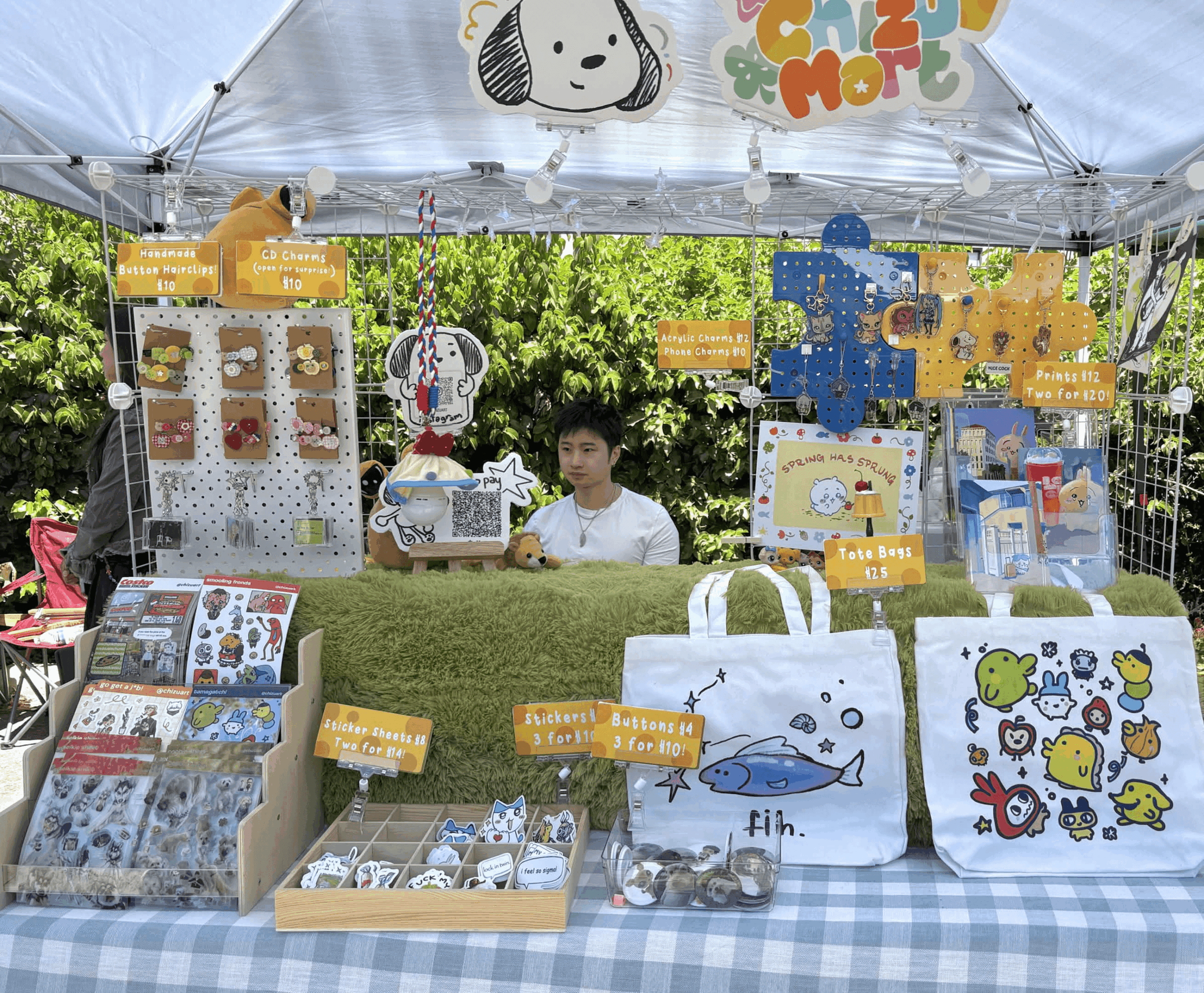 Person sitting at a booth with colorful plush toys, bags, and stationery items displayed outdoors under a canopy.