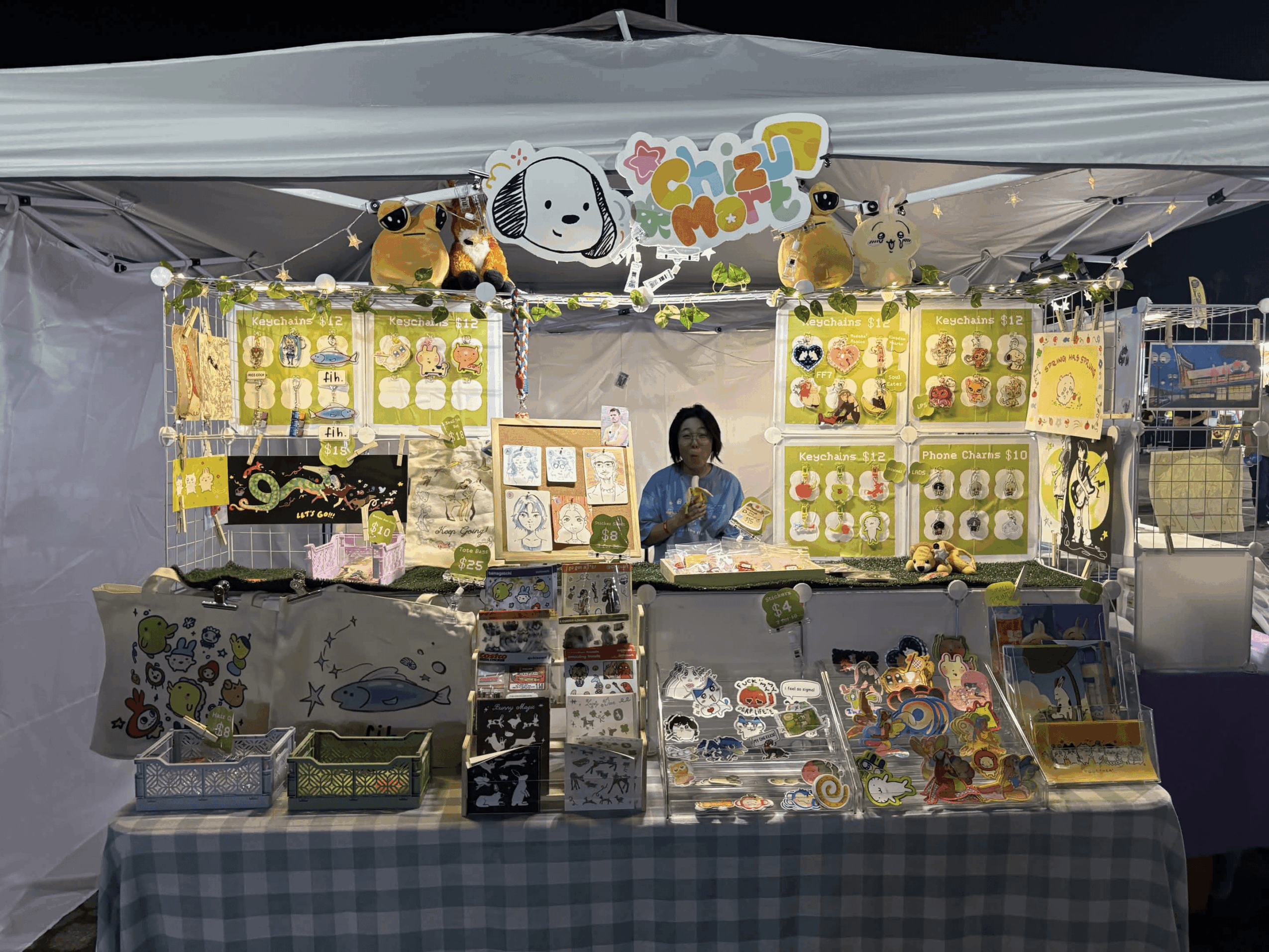 Vendor booth with plush toys, colorful displays, and a person behind the table at night, under a canopy with a sign.