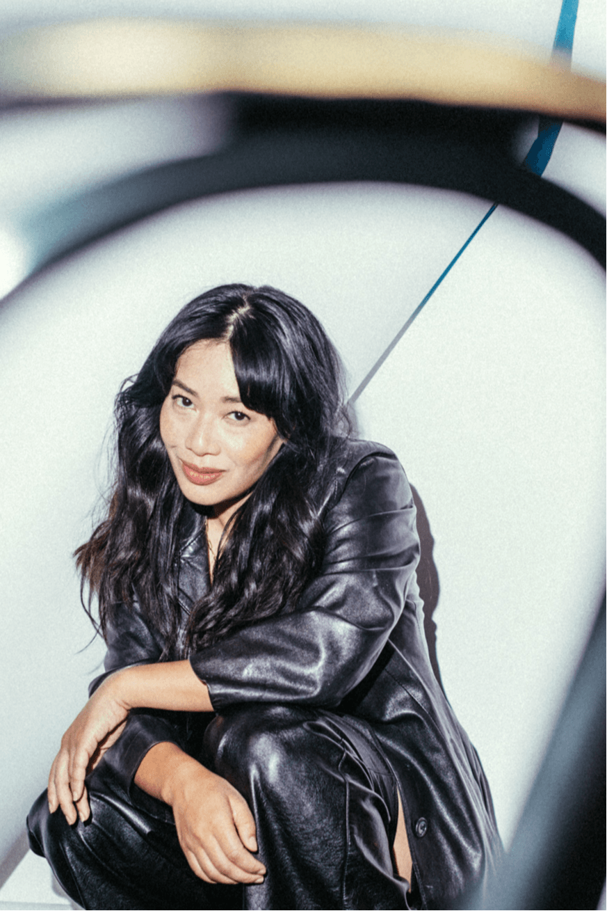 Woman with long dark hair wearing a black leather jacket, sitting with her hand on her knee, smiling.