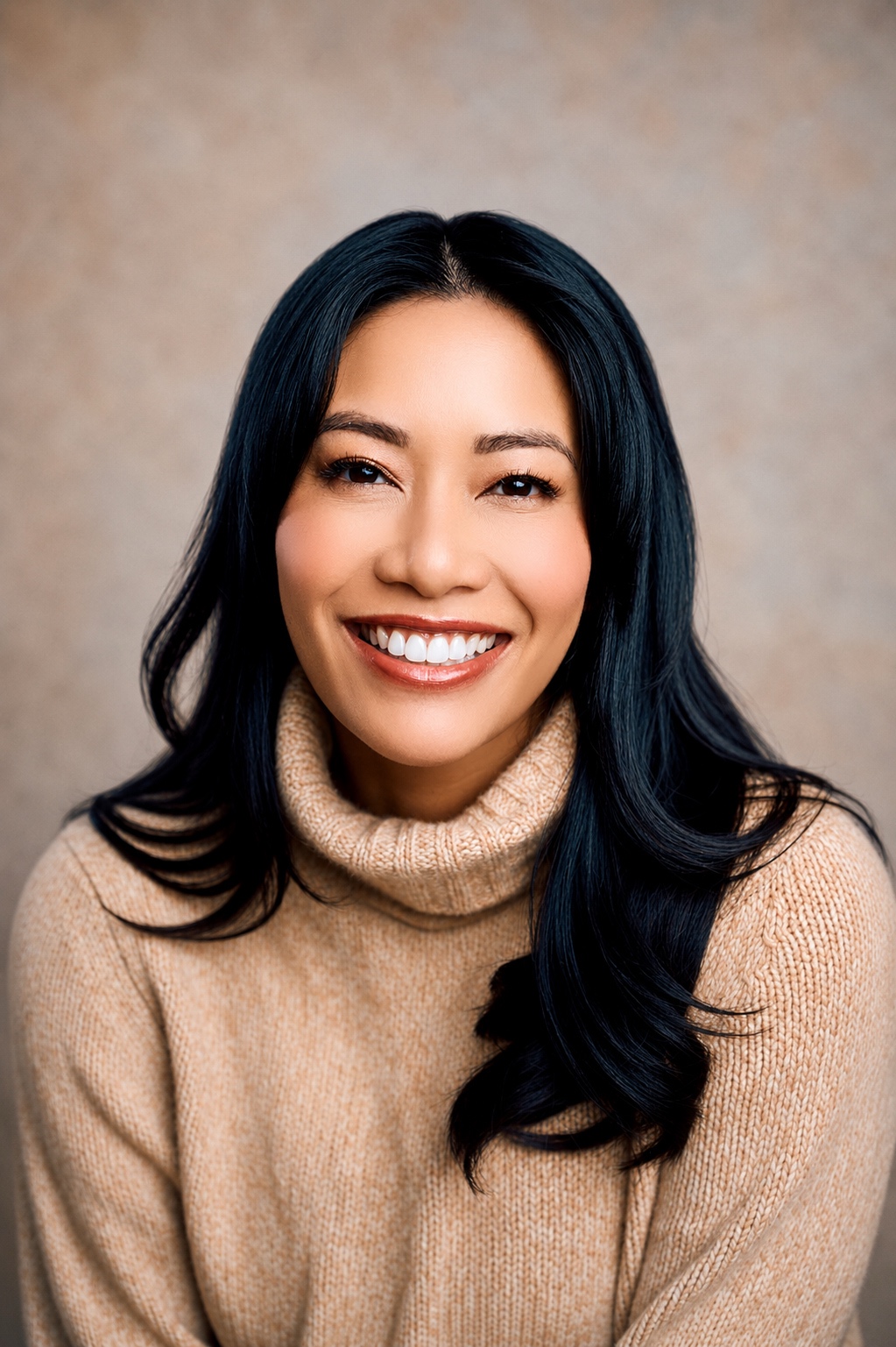 Smiling woman with long black hair wearing a beige turtleneck sweater, neutral background