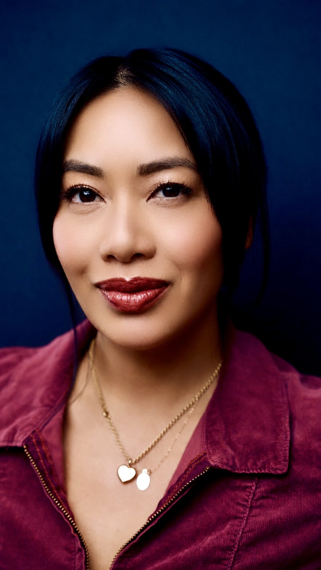 Portrait of a woman with dark hair, wearing a maroon jacket and gold necklace, smiling against a dark background.
