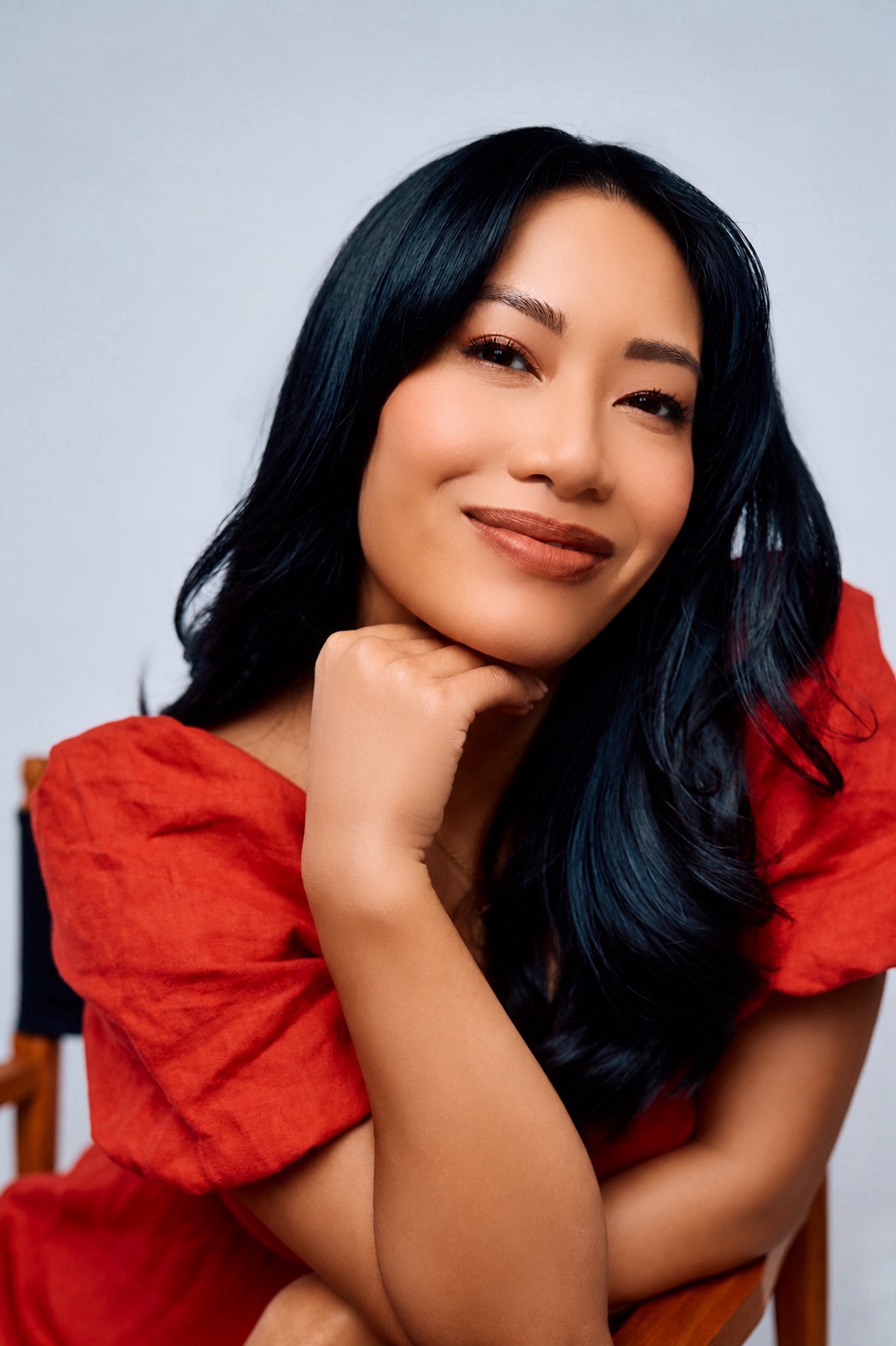 Woman with long black hair and a red top smiling, resting her chin on her hand, against a plain background.