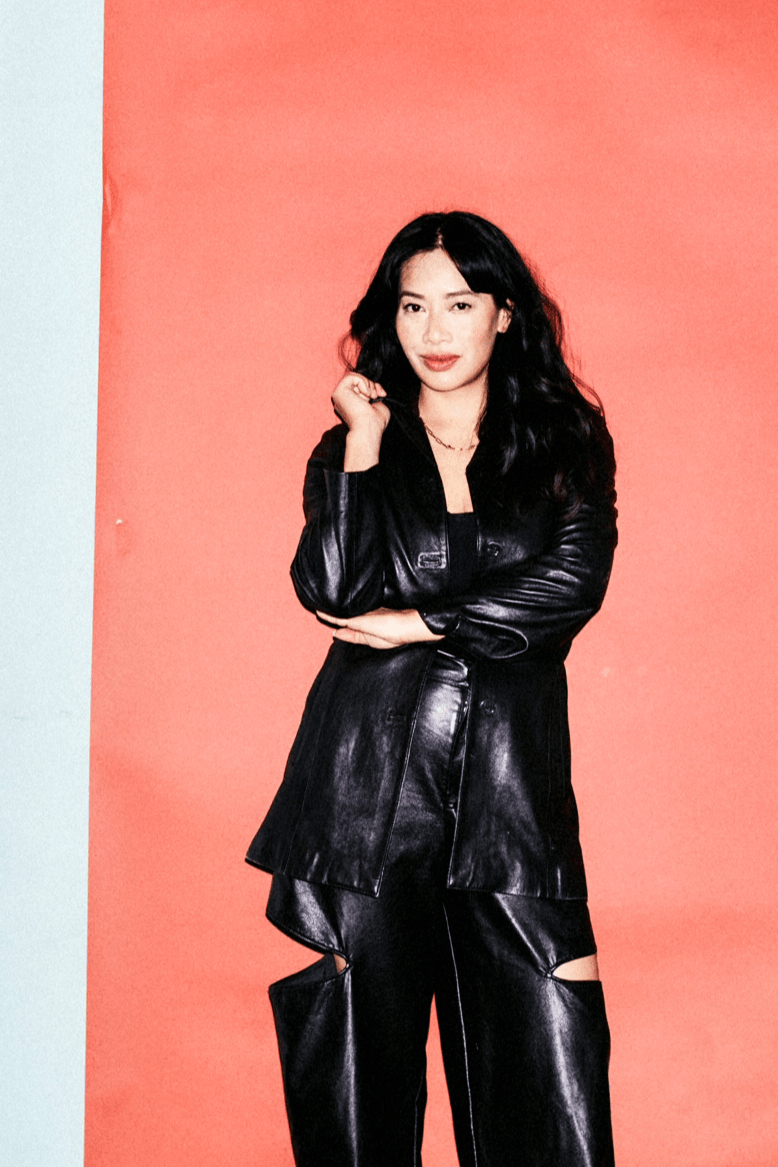 Woman with long black hair wearing a black leather outfit, standing against a pink background, smiling with her hand near her face.