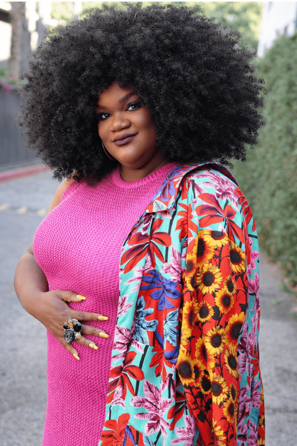 Woman with large curly hair wearing a pink top and colorful floral jacket, standing outdoors.