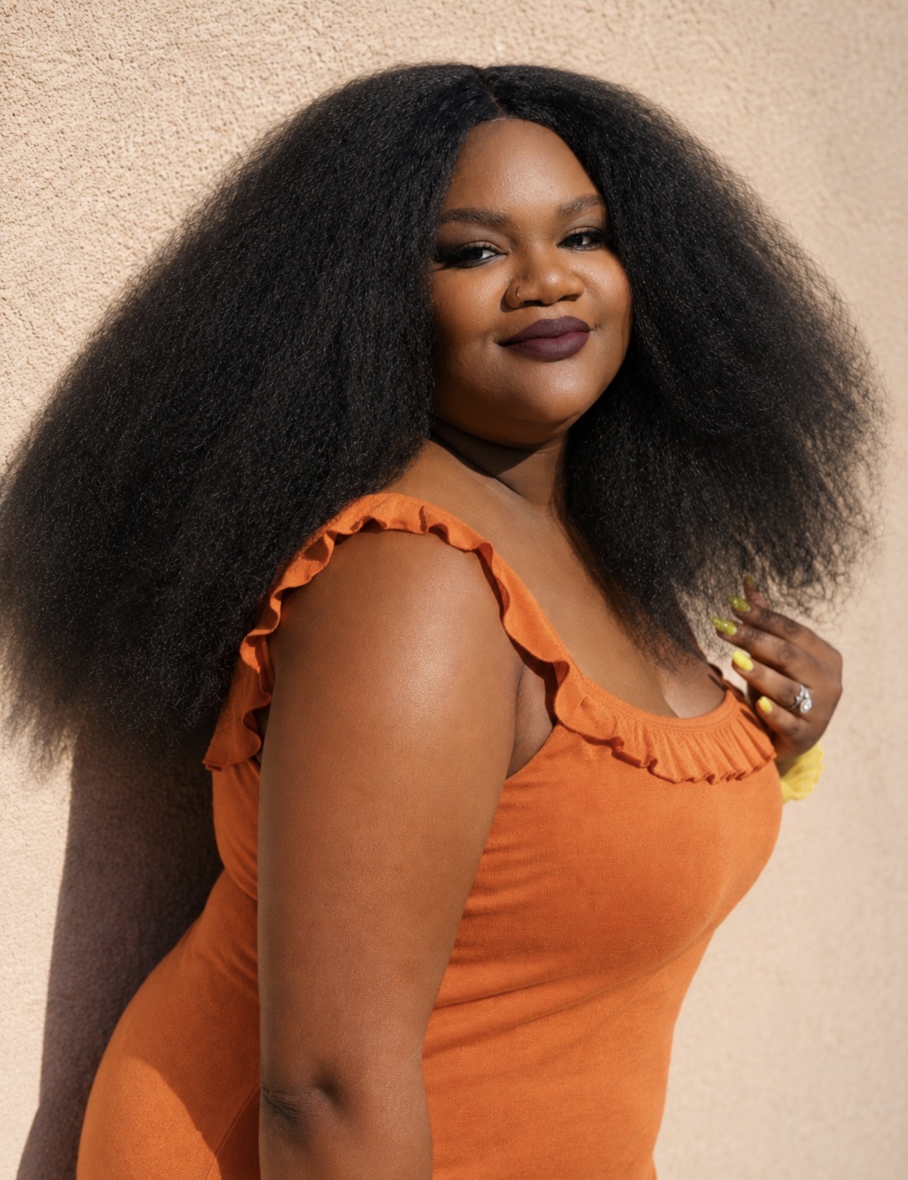 Woman with long, voluminous hair wearing an orange ruffled dress, smiling, against a neutral background.
