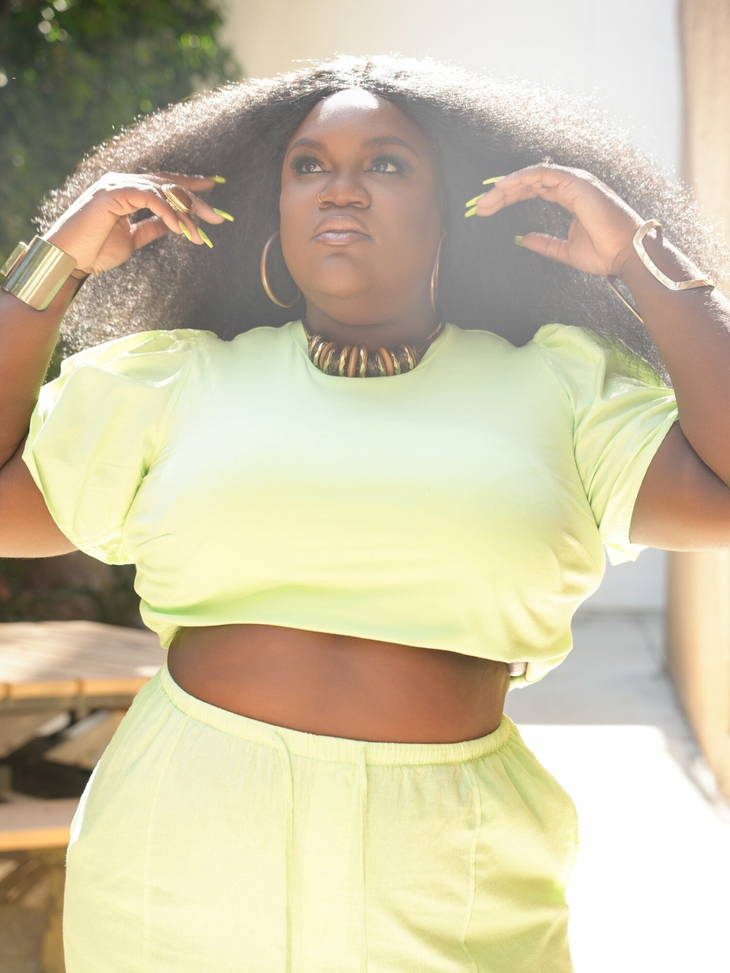 Woman with large curly hair wearing a yellow crop top and matching pants, outdoors with sunlight and greenery.