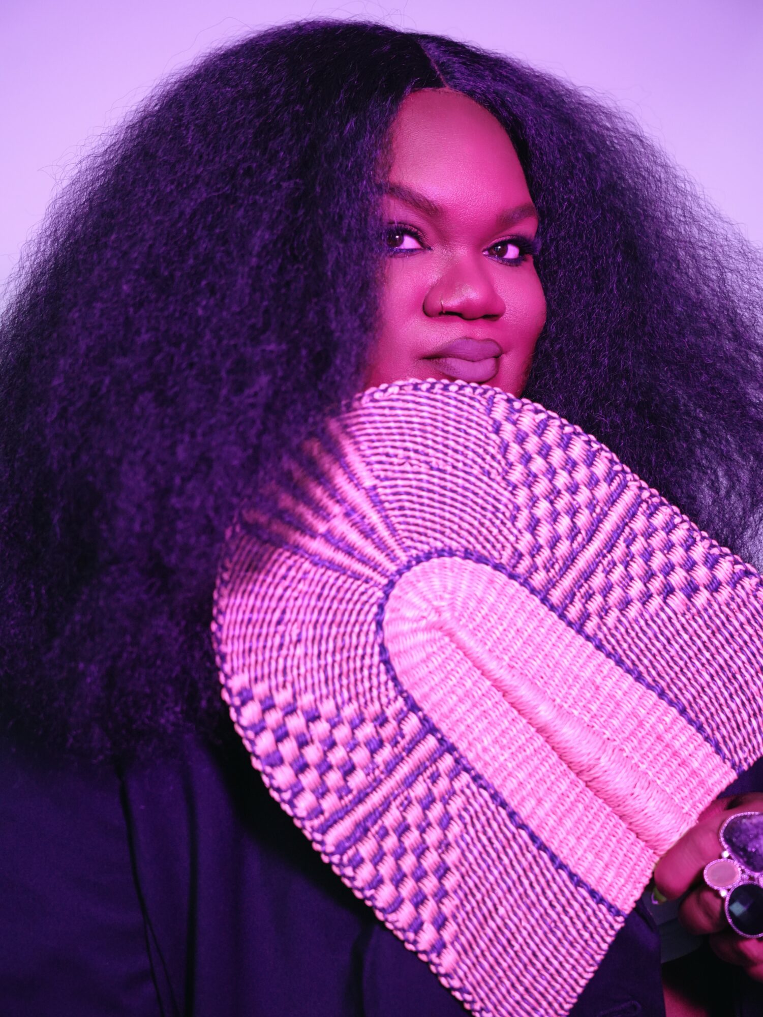 Woman with voluminous curly hair holding a patterned fan close to her face, looking at the camera, purple lighting.