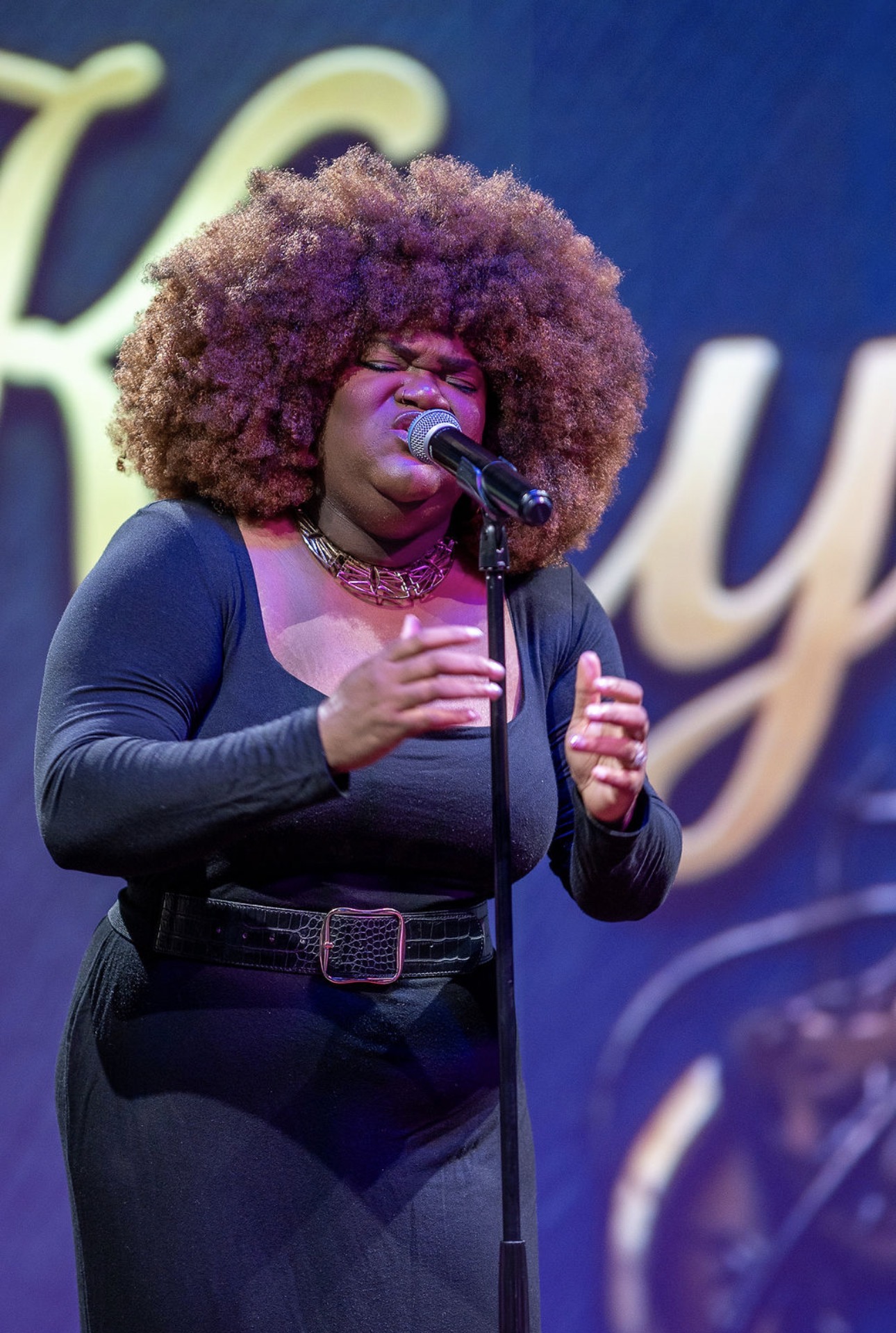 A woman with large curly hair sings into a microphone on stage, wearing a black outfit and a necklace.