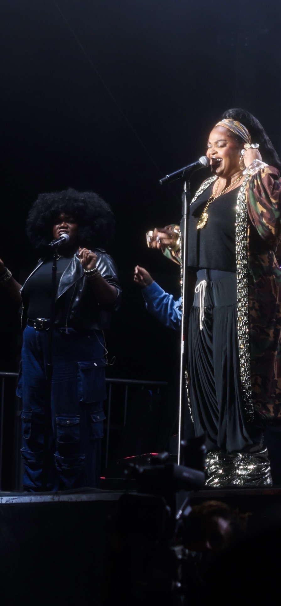 Two women on stage, one singing into a microphone, the other holding a small object, both dressed in dark clothing with stage lighting.