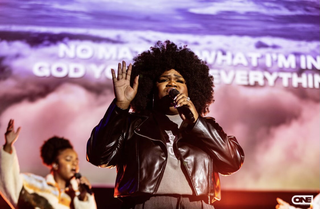 Woman with curly hair singing into a microphone on stage, with a background of clouds and text, and a child in the background.