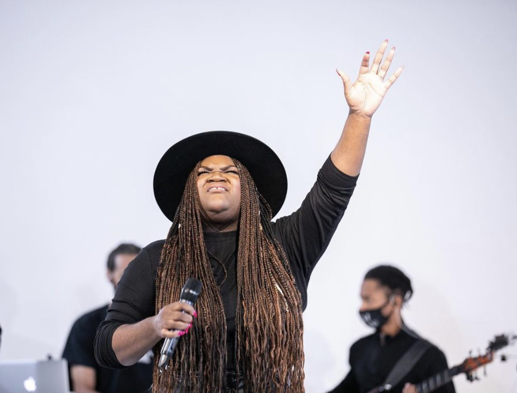 Woman with long hair and a hat raising her hand, holding a microphone, with musicians in the background.