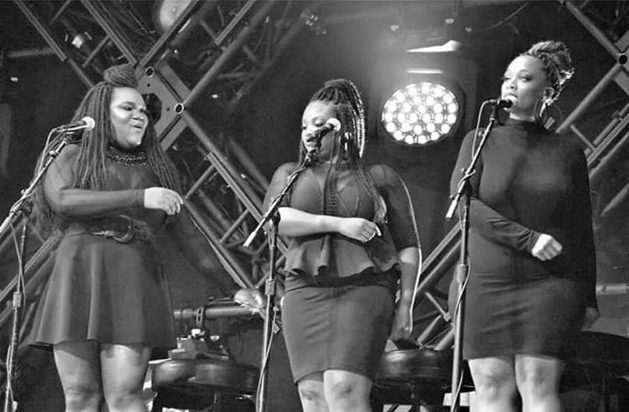 Three women singing on stage with microphones, stage lights, and metal framework in background.