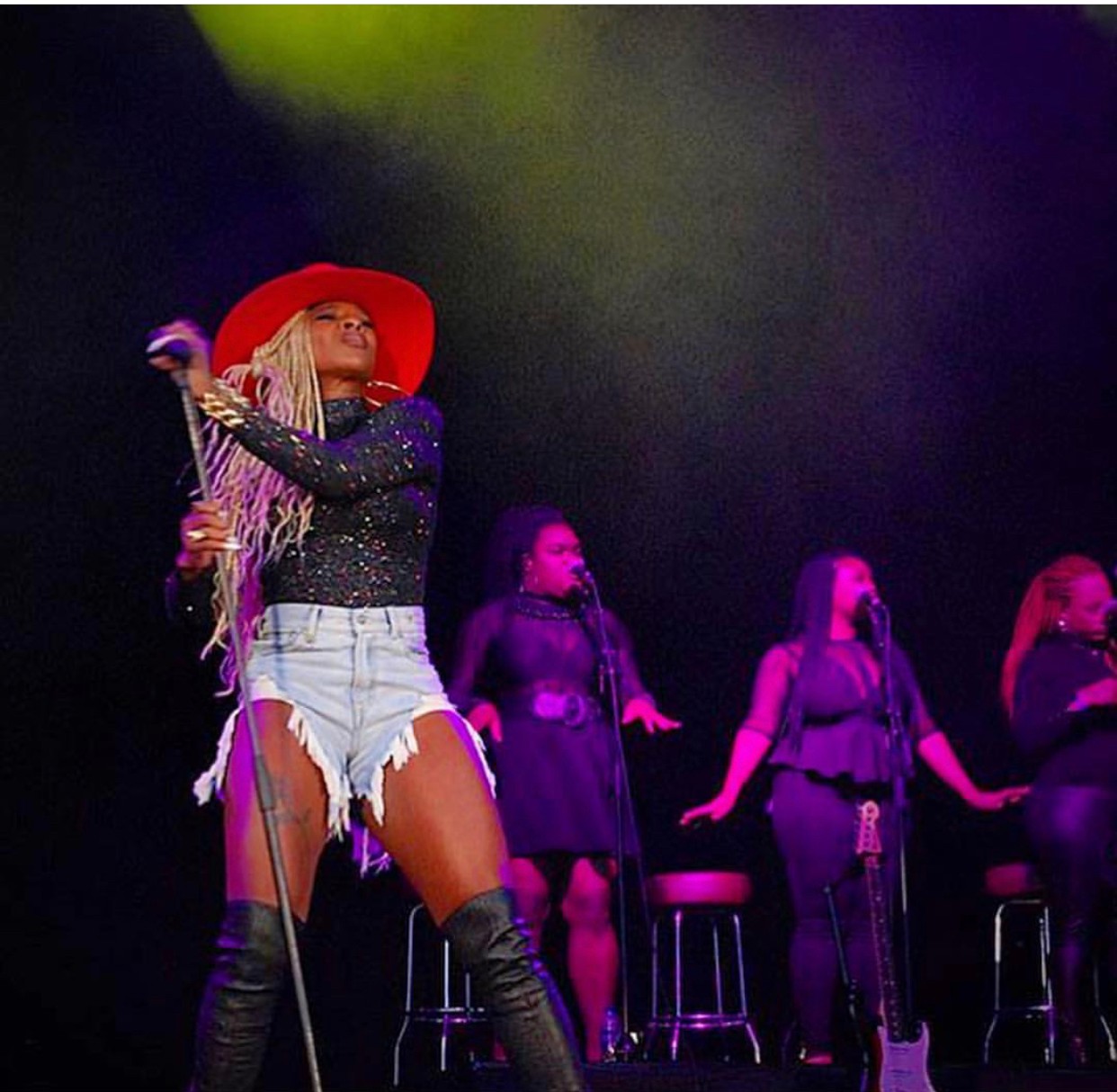 Performer with a red hat singing on stage, three backup singers in black outfits behind her, colorful stage lighting.