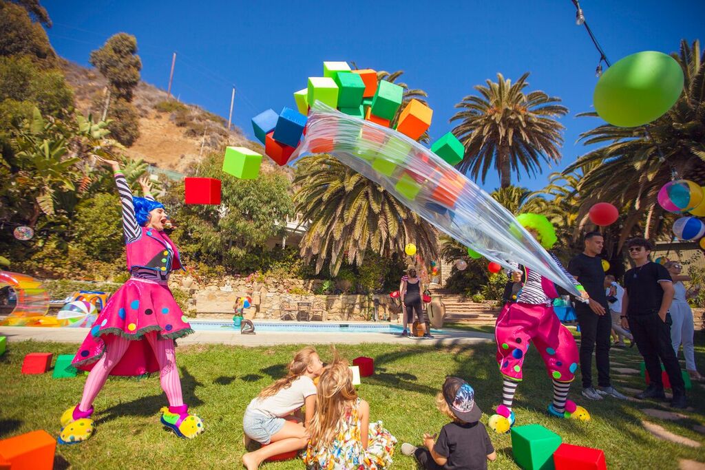 Children playing with colorful balloons and clowns in a park with trees and palm trees in the background.
