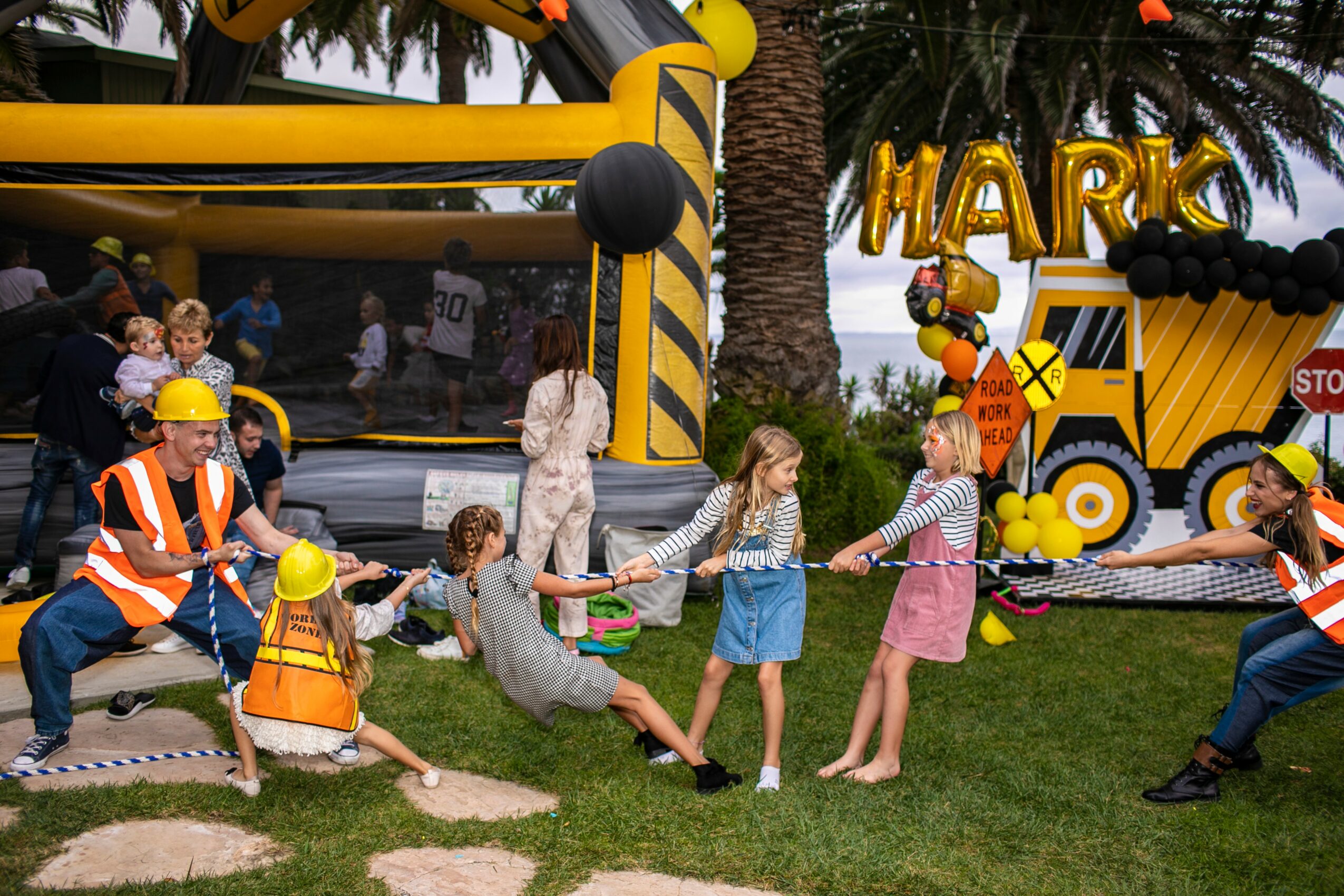 Children and adults playing tug-of-war outdoors near a bounce house decorated with balloons, with trees and grass in the background.