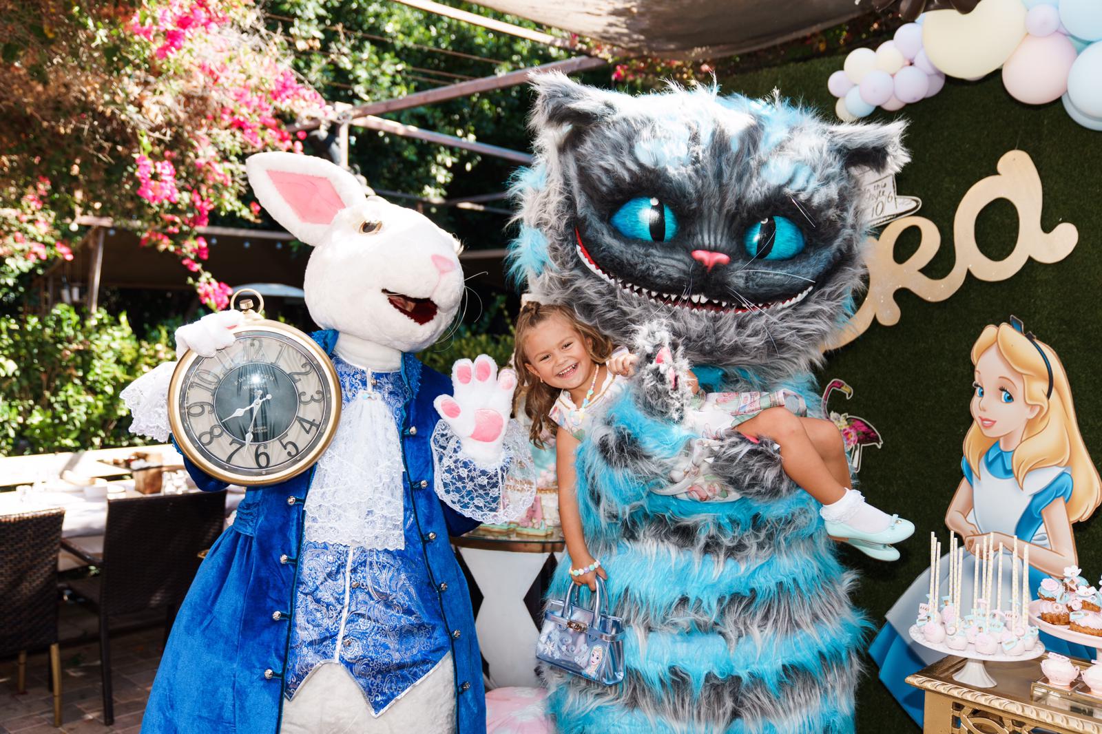 Child with two costumed characters, one resembling a white rabbit with a clock, and a large blue striped cat, outdoors with pink flowers and balloons