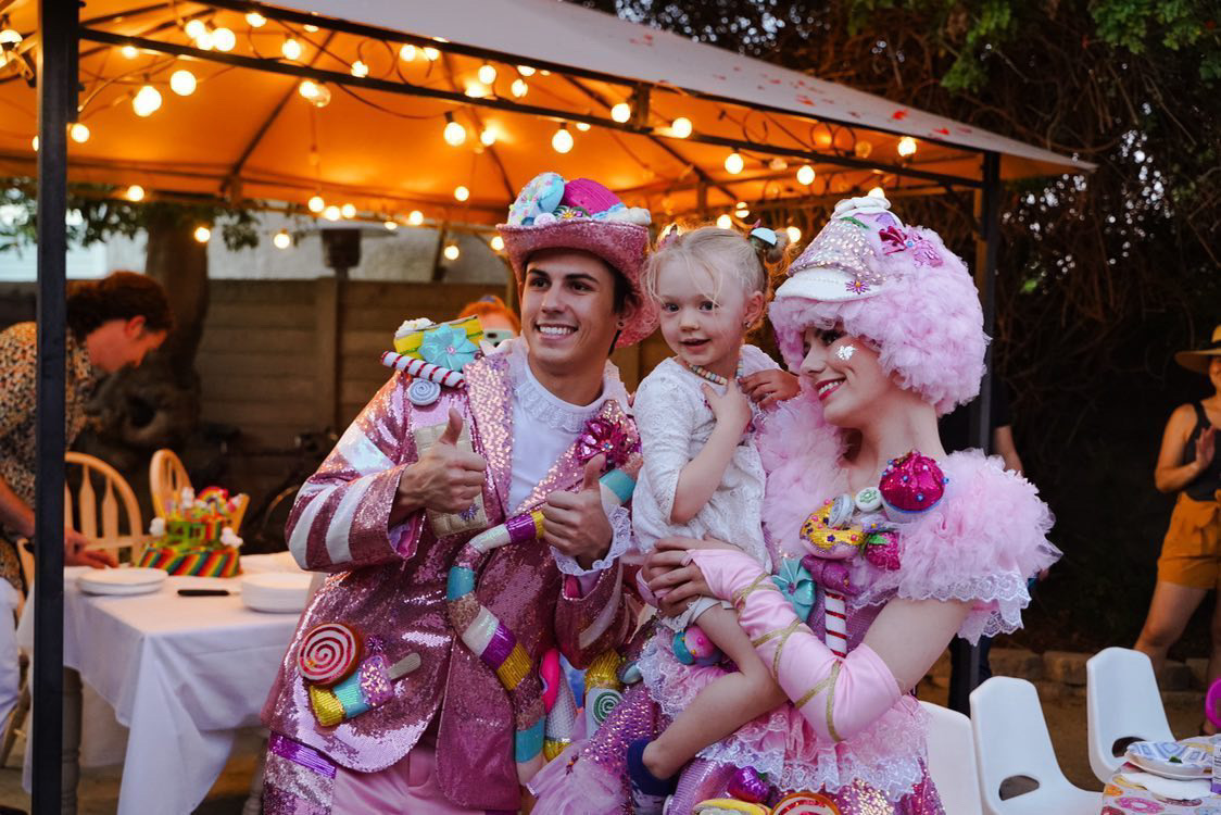 Two performers in colorful costumes with children at an outdoor event, decorated with lights and tables, during evening.