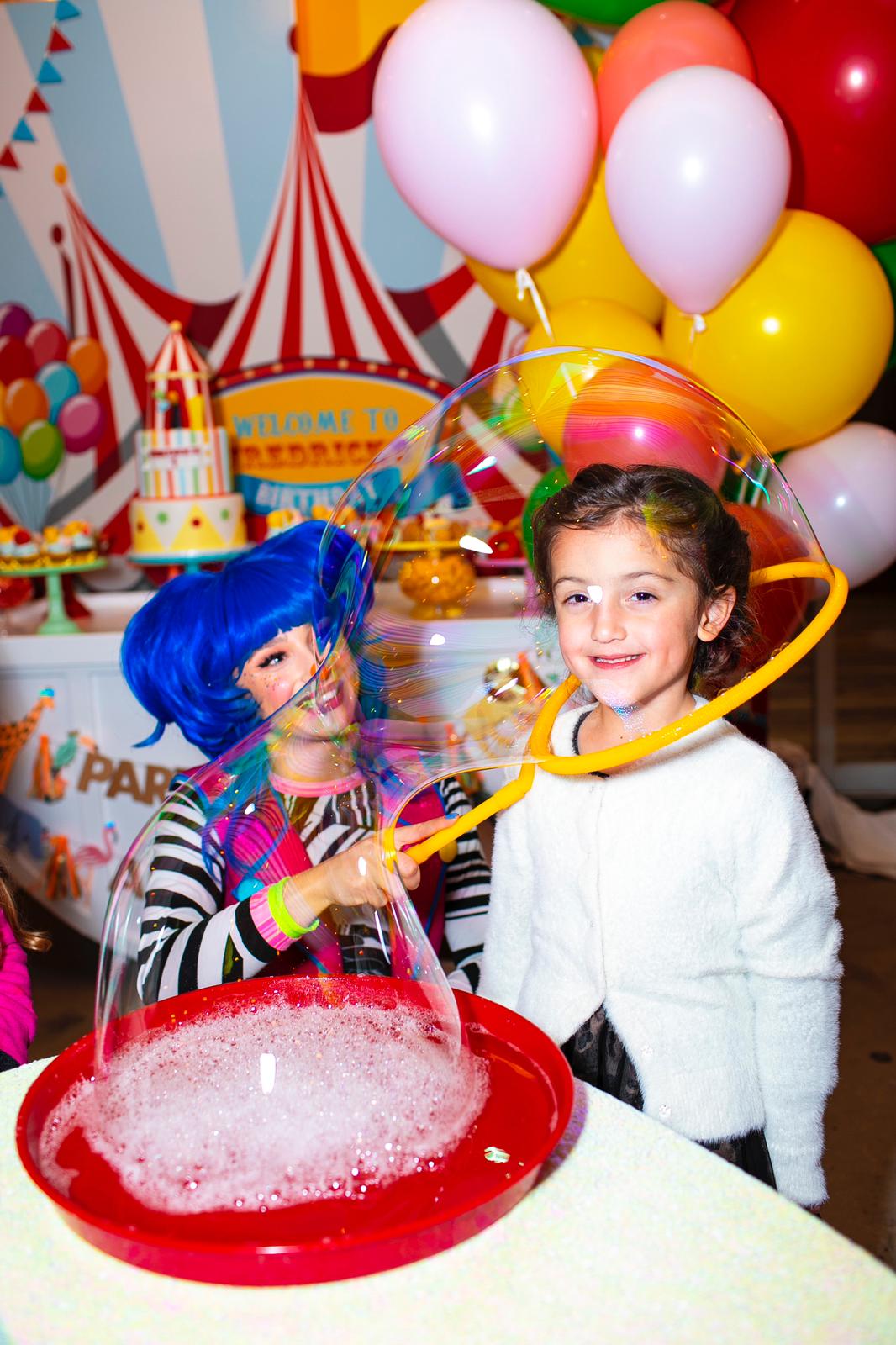 Children at a birthday party with balloons, a cake, and colorful decorations, one girl smiling and another in costume.