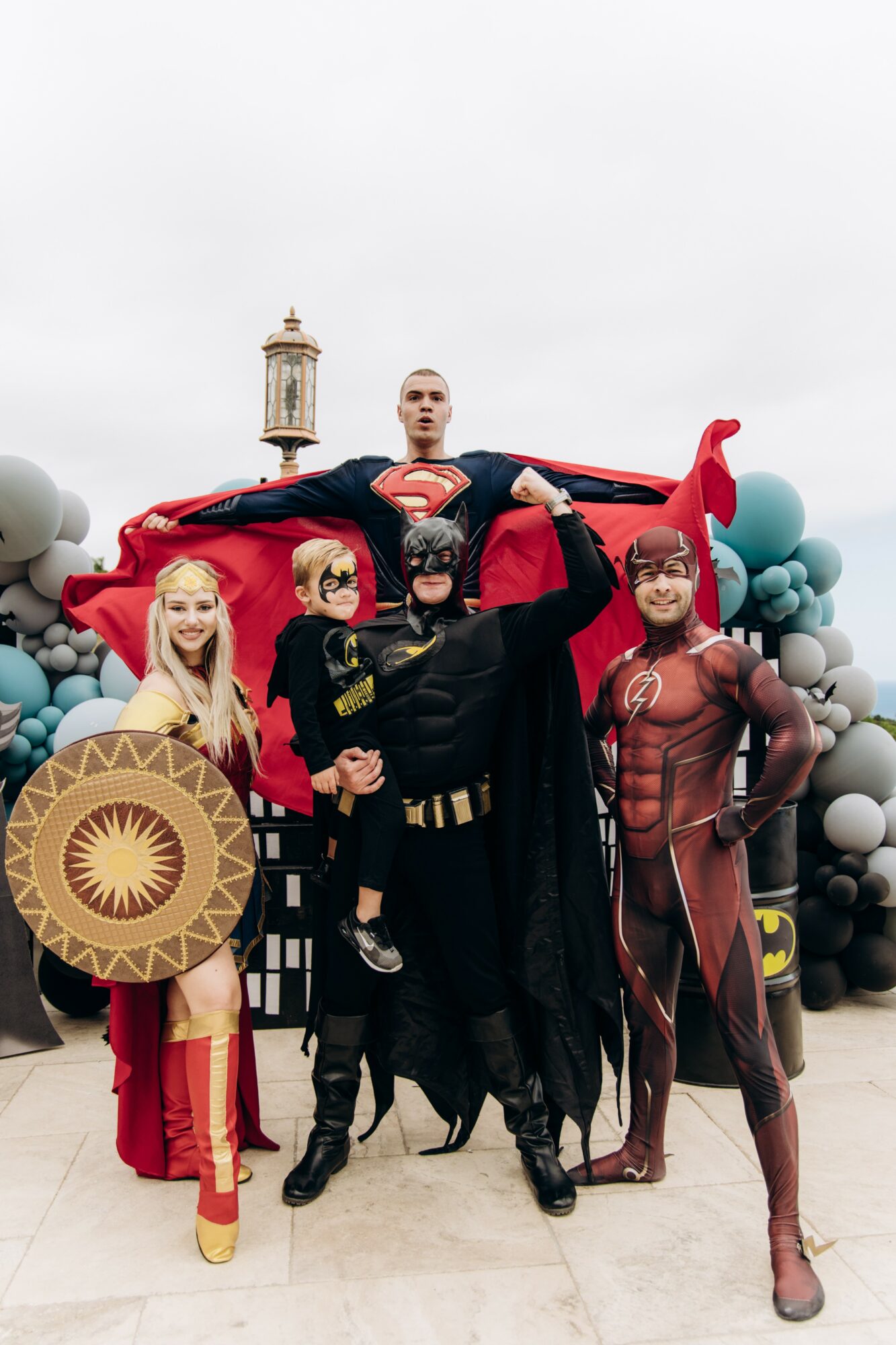 Group of people dressed as superheroes, with a person in a Superman costume lying on a red fabric, outdoors with cloudy sky and a tower in background.